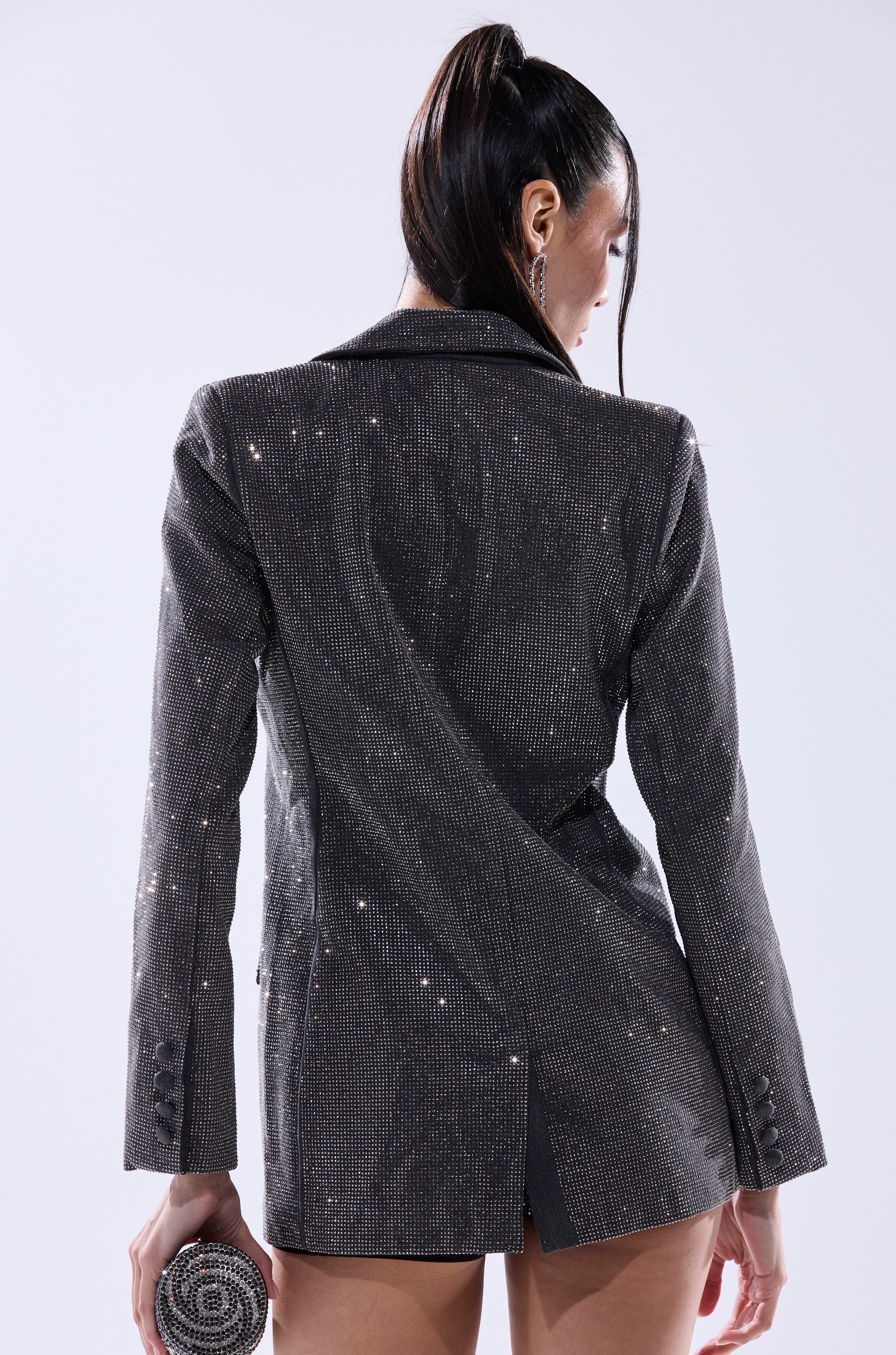 A woman with long, dark hair in a high ponytail is seen from behind wearing the ALL EYES ON ME EMBELLISHED DENIM BLAZER and holding a round, glittery clutch against a plain light gray background. 5