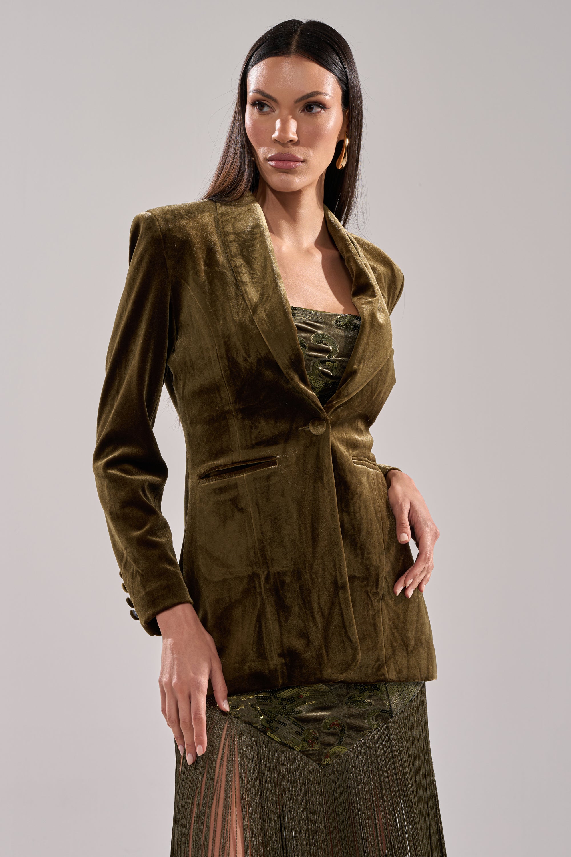 A woman wearing the DIRTY MARTINI VELVET BLAZER over a patterned green dress stands against a plain background, gazing to the side. She has long, straight dark hair and gold hoop earrings.