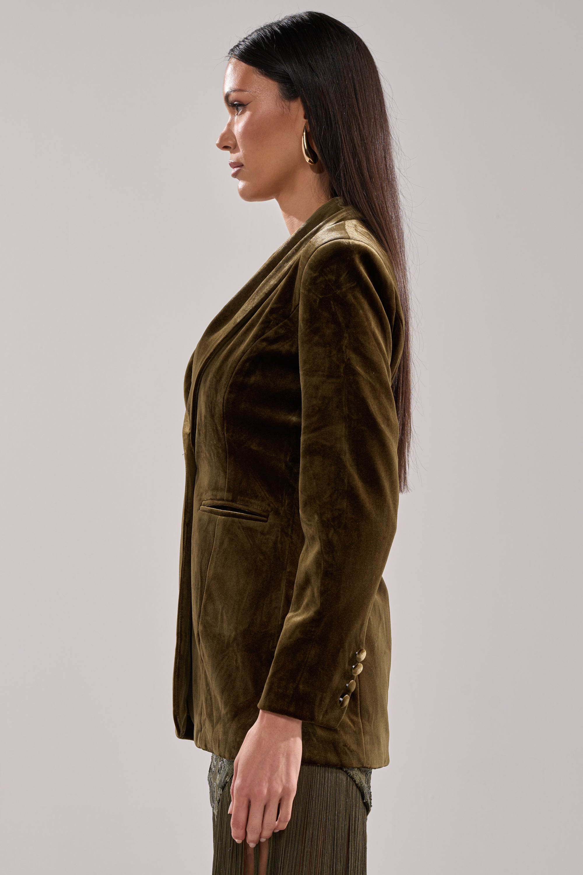 A woman with long straight hair models the DIRTY MARTINI VELVET BLAZER in brown and gold hoop earrings, standing in profile against a light gray background.