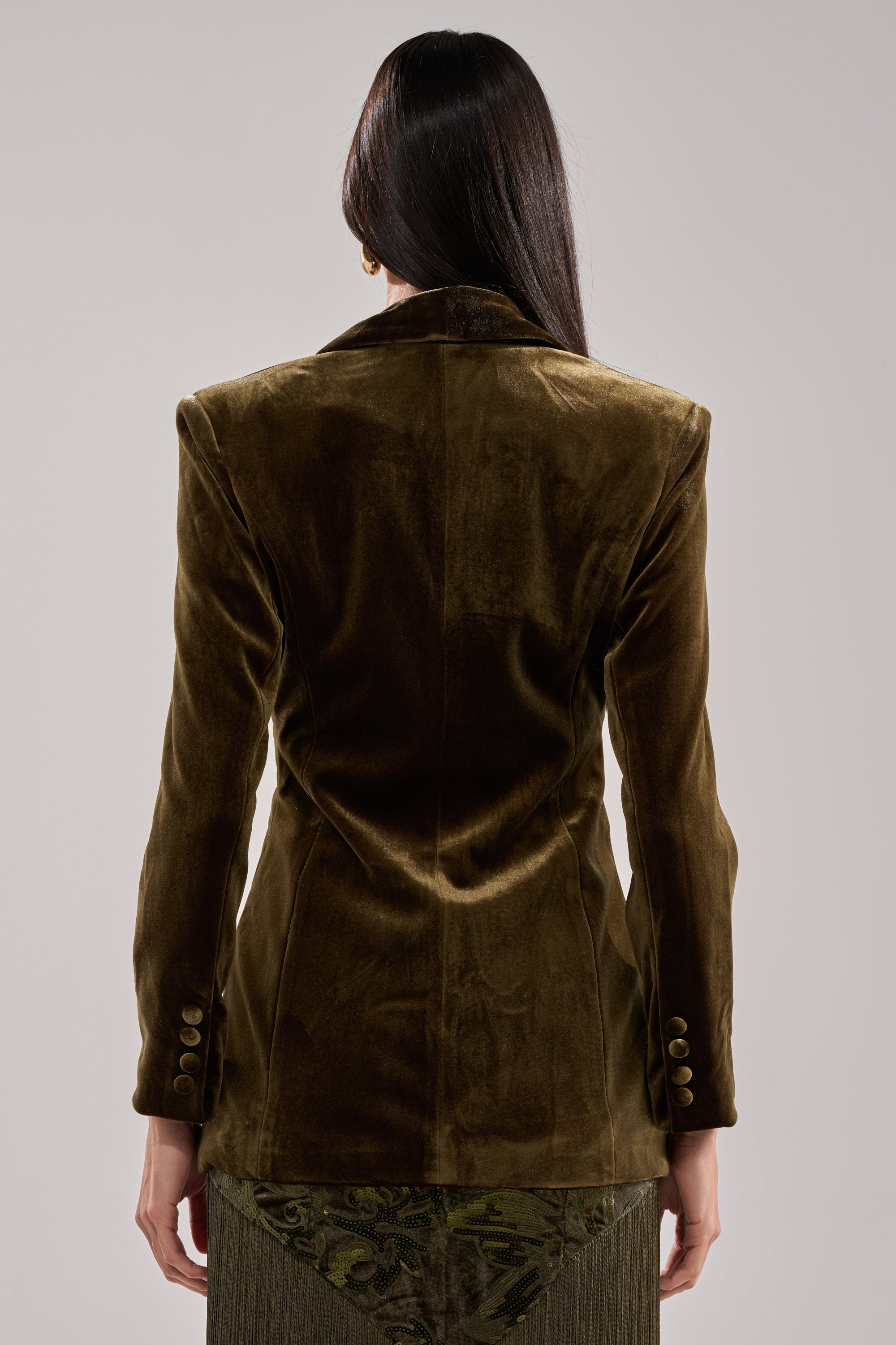 A person with long dark hair is seen from the back, wearing the DIRTY MARTINI VELVET BLAZER in olive green, featuring sleeve button details and a patterned green skirt peeking out below.