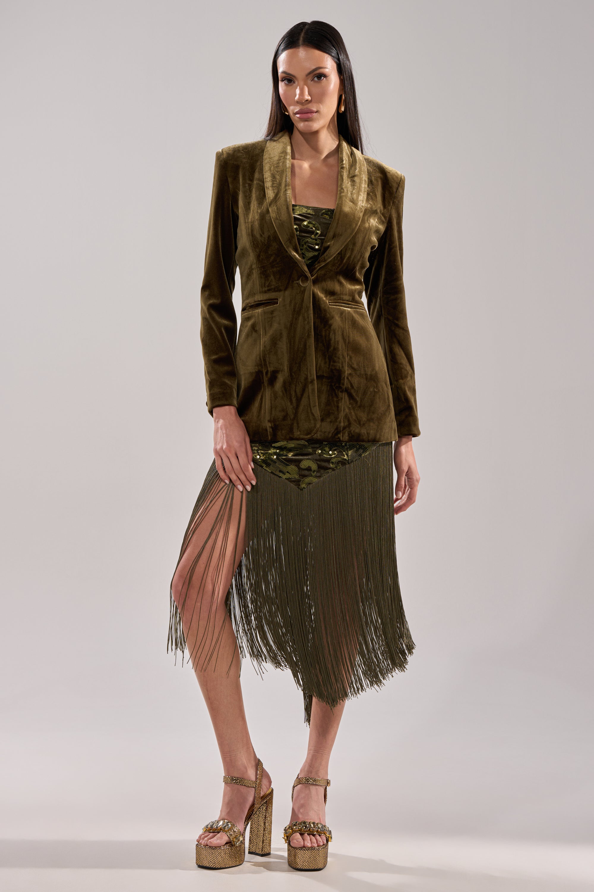 A woman poses against a light gray background wearing the DIRTY MARTINI VELVET BLAZER in brown over a green fringe dress and gold platform heels. She has long dark hair and a neutral expression.
