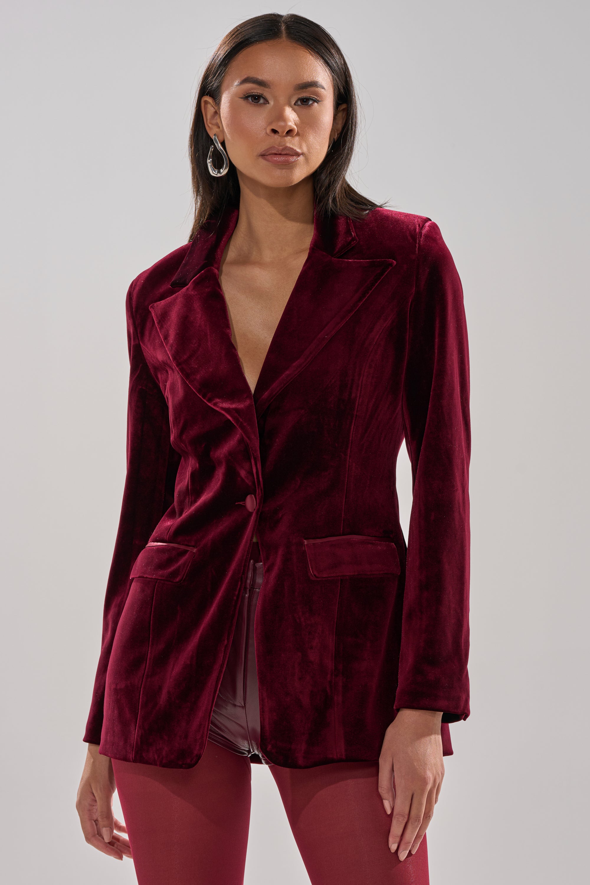 A woman with long dark hair and hoop earrings poses confidently in the LUXE VELVET TUXEDO BLAZER, a red single-button piece paired with matching pants, putting a modern spin on the classic tailored suit against a plain background.