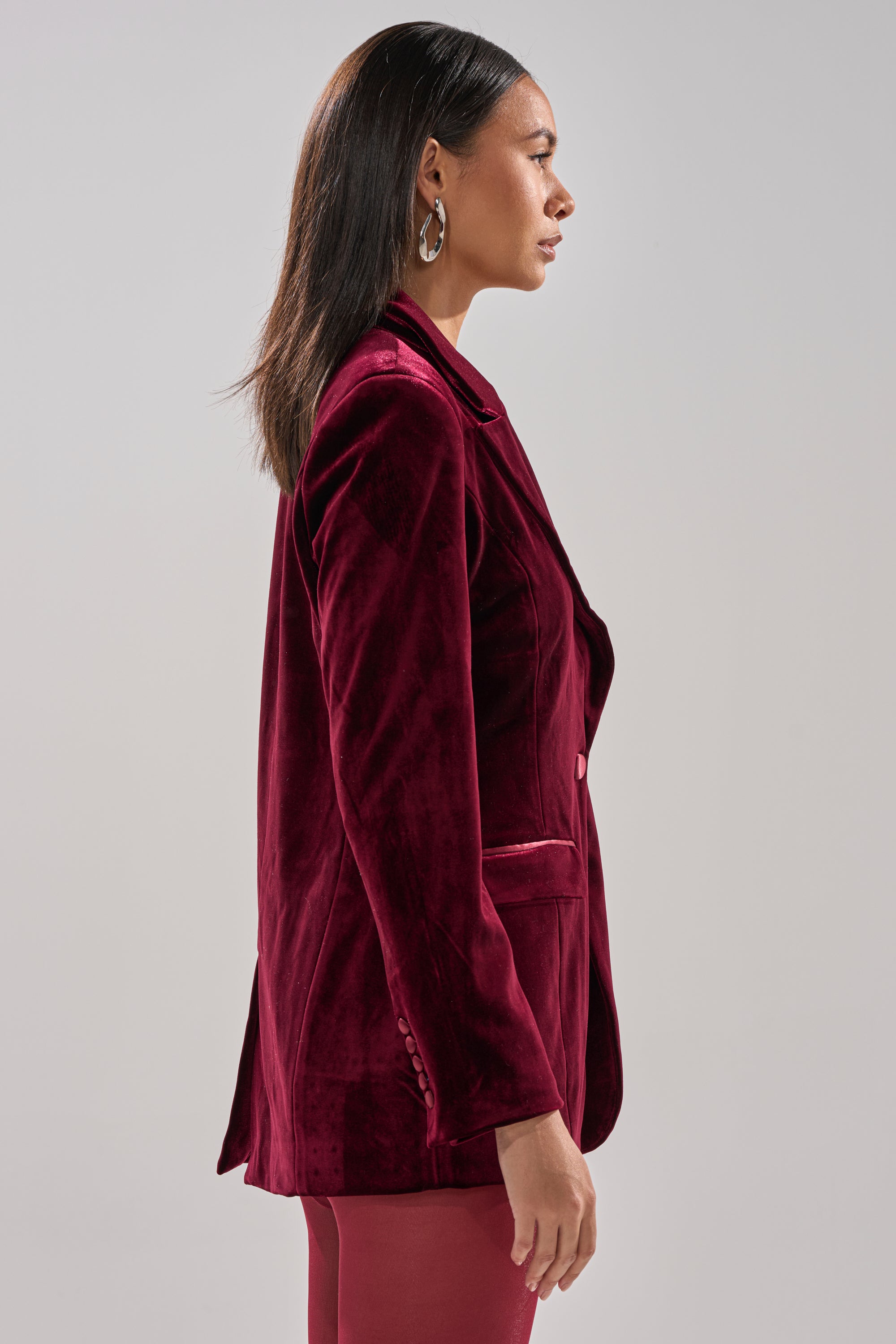A woman with straight dark hair, in large hoop earrings and the LUXE VELVET TUXEDO BLAZER, stands in profile against a plain light background.