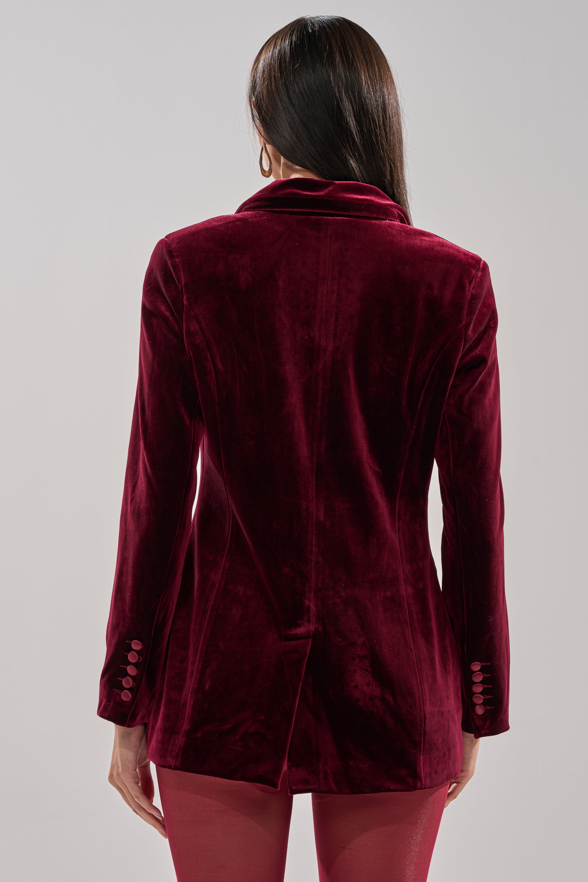 A woman with long dark hair is seen from behind, wearing the LUXE VELVET TUXEDO BLAZER with button-detailed sleeves and matching red pants, set against a plain light background.