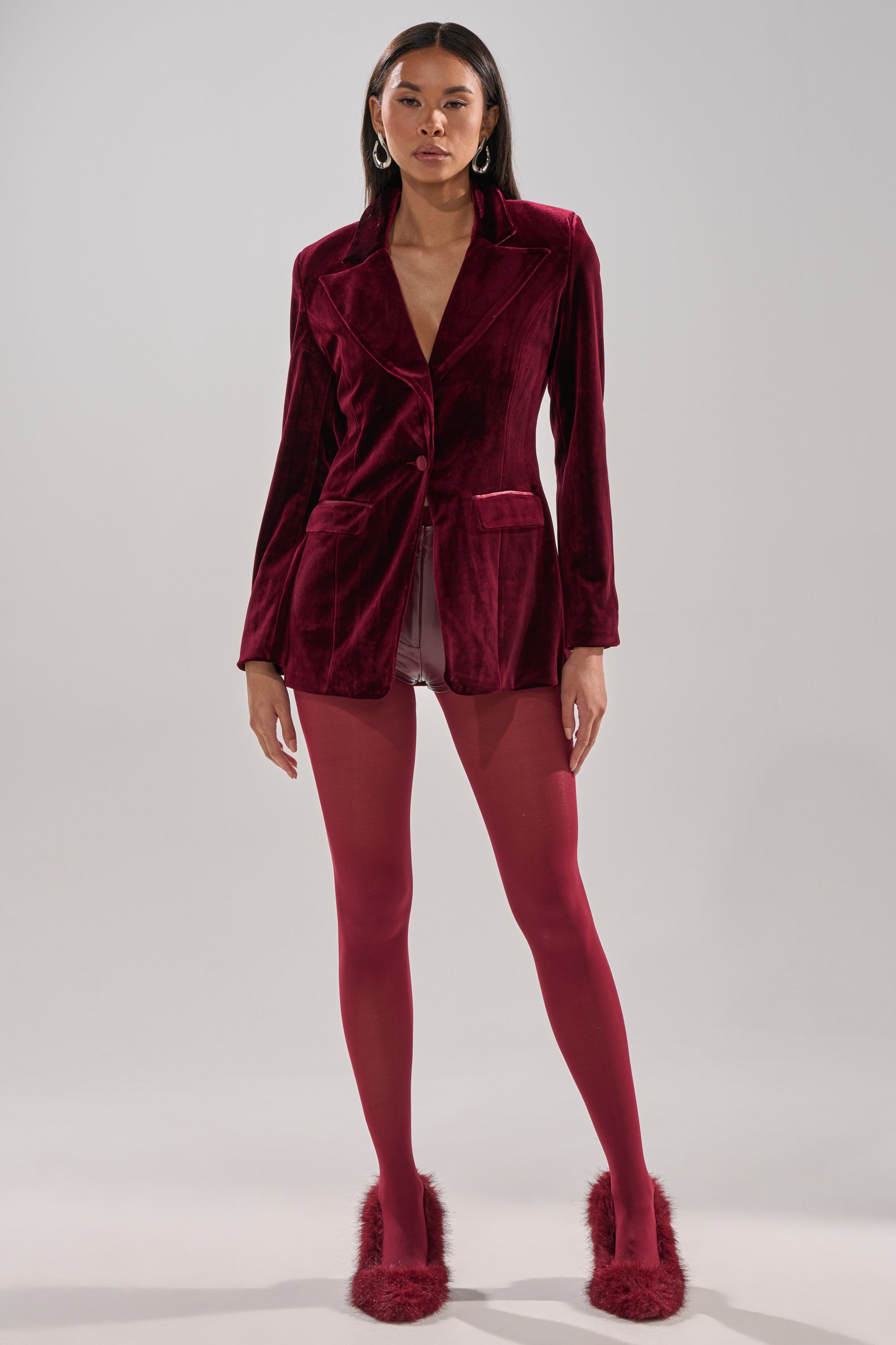 A woman faces forward against a light background, wearing the LUXE VELVET TUXEDO BLAZER in red, paired with matching red tights, light satin shorts, large hoop earrings, and red furry-heeled shoes.