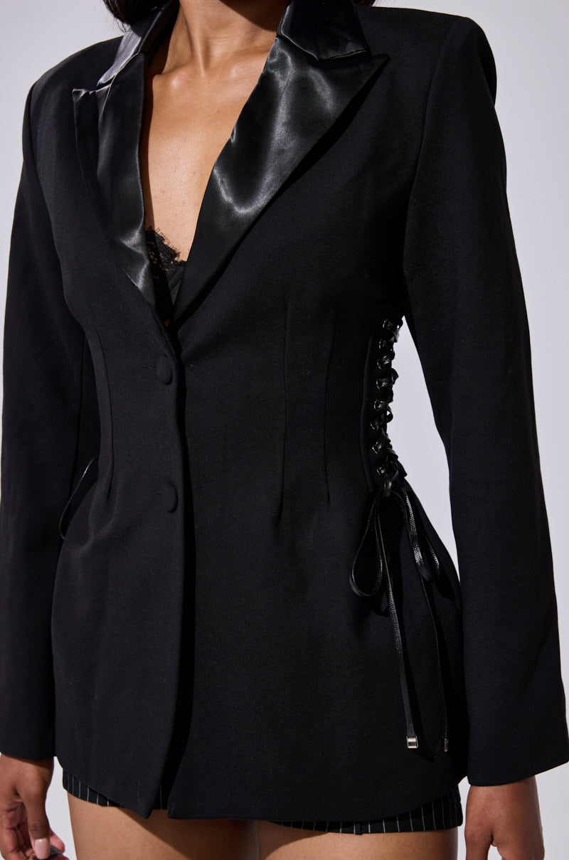 A woman models the SCARLET CORSETED BLAZER with satin lapel and lace-up side panels over a lace top; only her torso and upper thighs are shown. 11