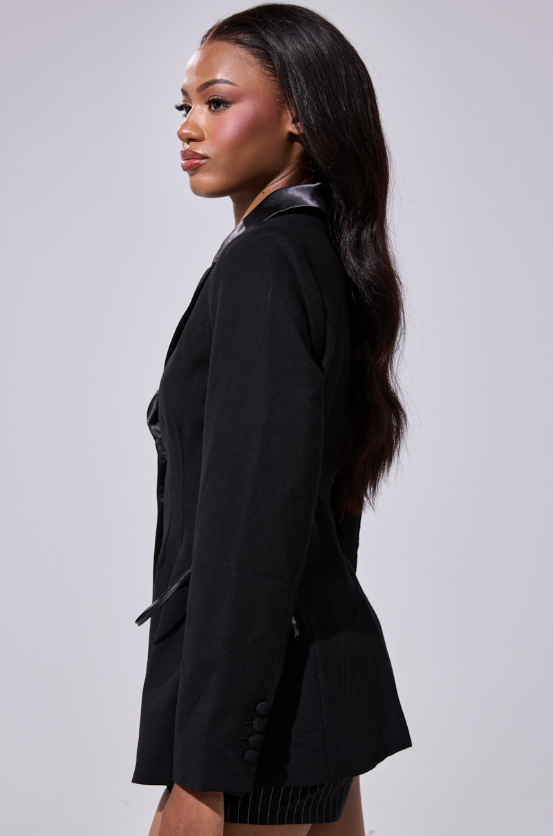 A woman with long, wavy hair stands in profile against a plain, light background, wearing the SCARLET CORSETED BLAZER paired with a pinstriped skirt. 6