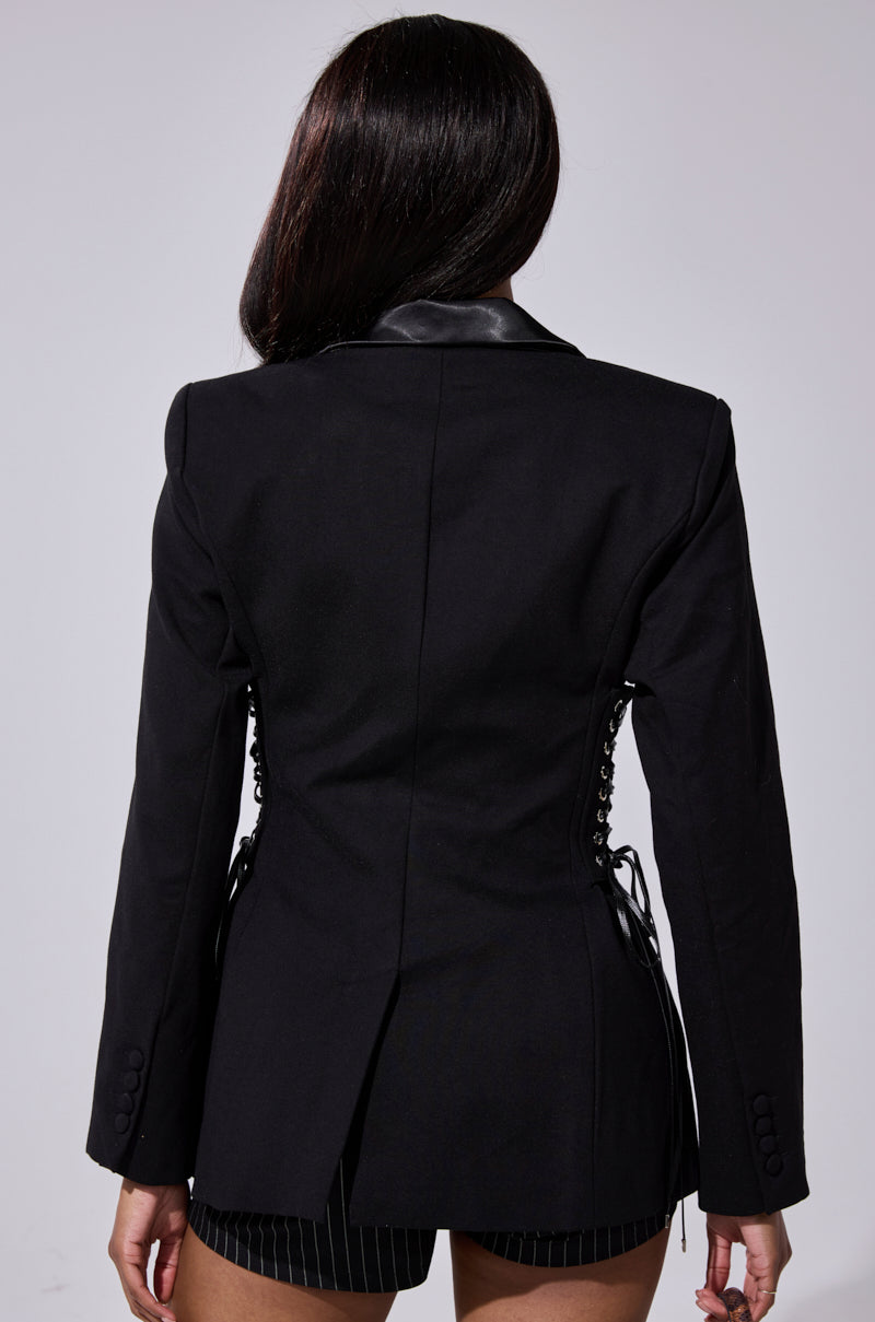 A person with long dark hair is seen from the back wearing the SCARLET CORSETED BLAZER, featuring a leather collar, lace-up sides, and paired stylishly with black pinstriped shorts. 7