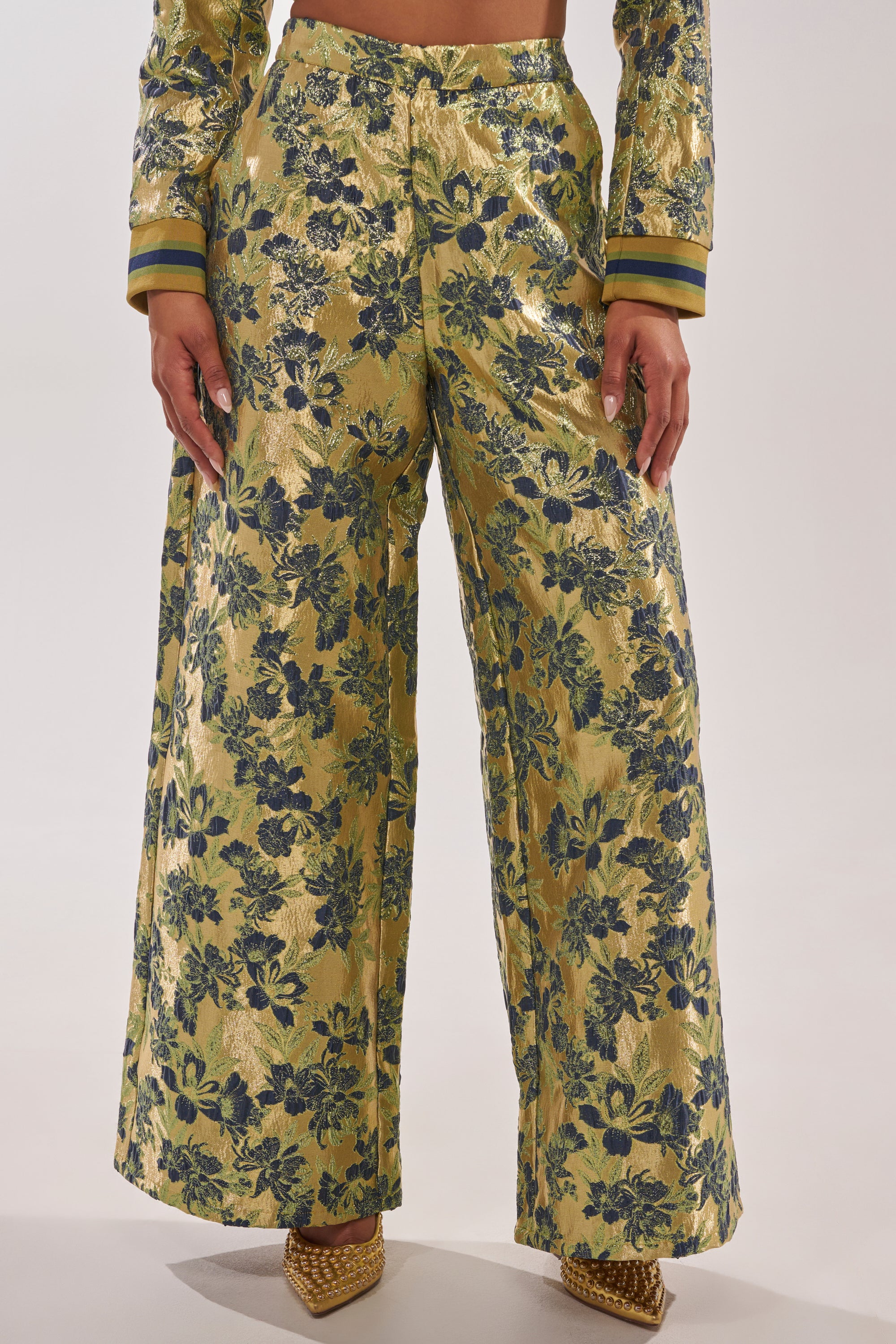 A person wears the HARLOW BROCADE WIDE LEG PANT with a blue and green floral pattern, matching jacket sleeves, and gold textured pointed-toe shoes against a plain light background. 1