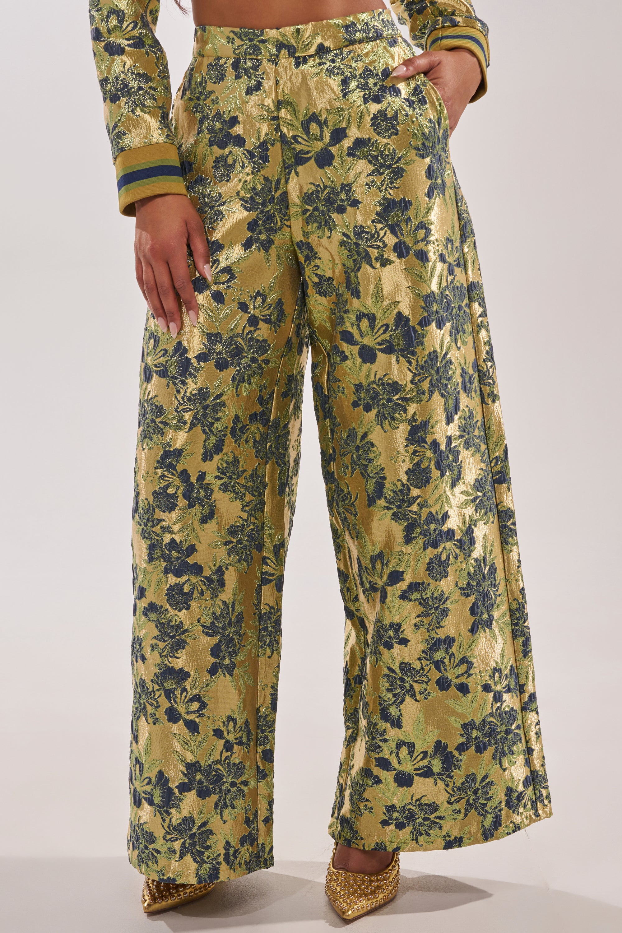 A person wears the HARLOW BROCADE WIDE LEG PANT, featuring a shiny blue and green floral pattern, matched with a coordinating top and gold pointed shoes. The photo is cropped at the chest and ankles. 2
