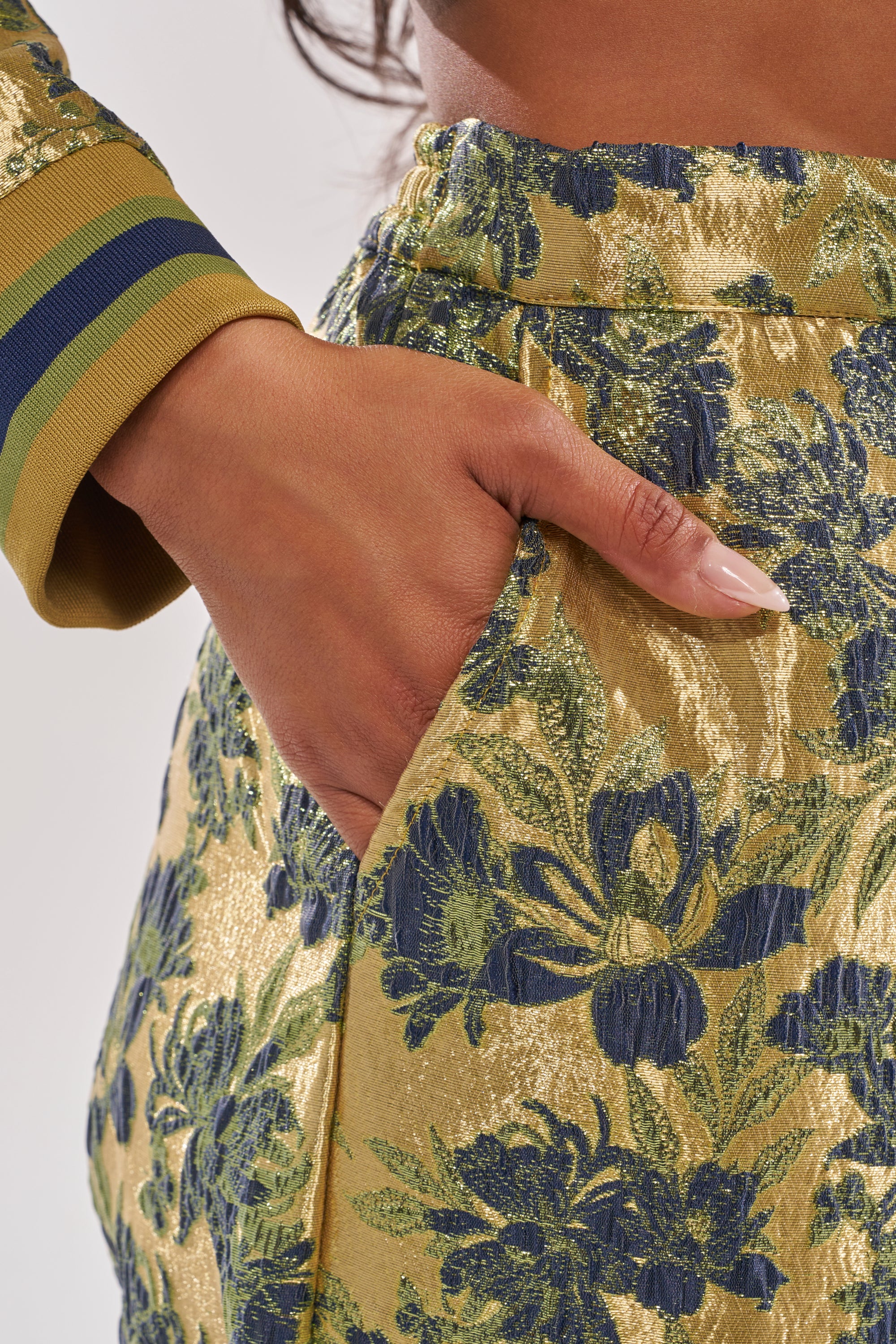 A close-up of a hand in the pocket of the HARLOW BROCADE WIDE LEG PANT, featuring a blue and green floral metallic brocade pattern, with part of a matching sleeve visible. 6
