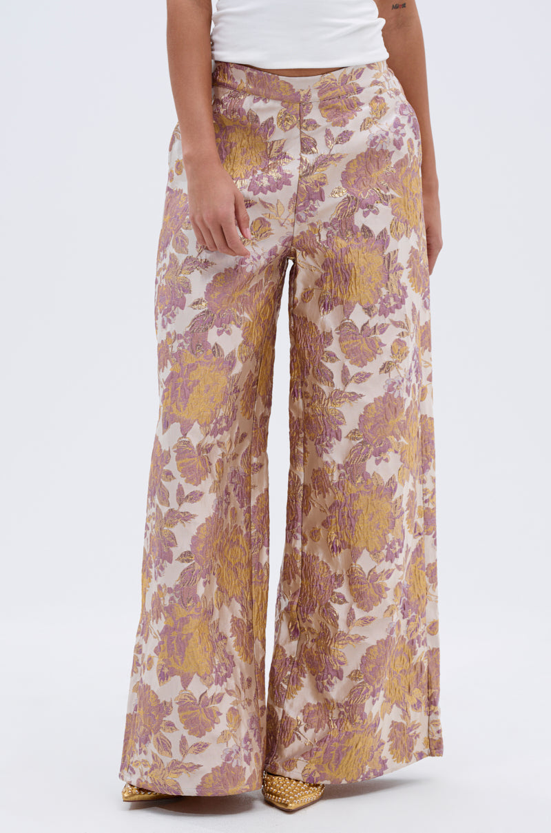 Wearing the SLOW MOTION BROCADE TROUSER featuring a pink and gold floral pattern, a person pairs them with a white top and yellow textured shoes, standing against a plain white background. 1