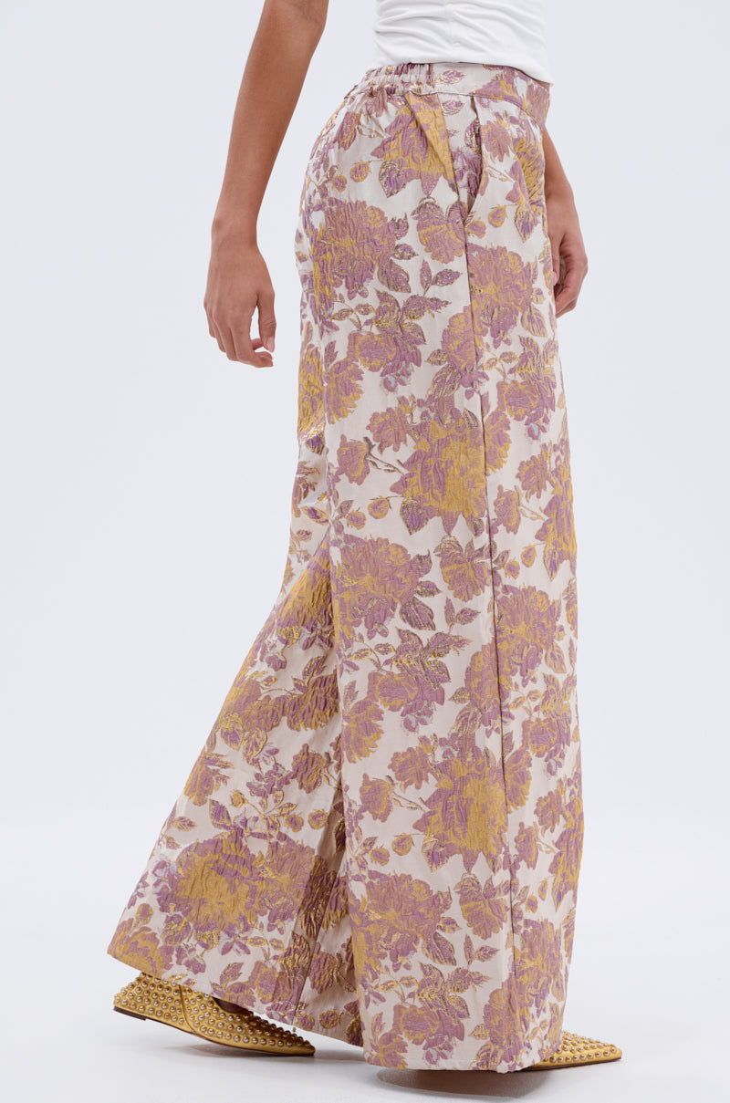 Someone is shown from the side against a plain white background, wearing SLOW MOTION BROCADE TROUSER with a pink and yellow floral pattern, paired with a white top and woven yellow shoes. 3