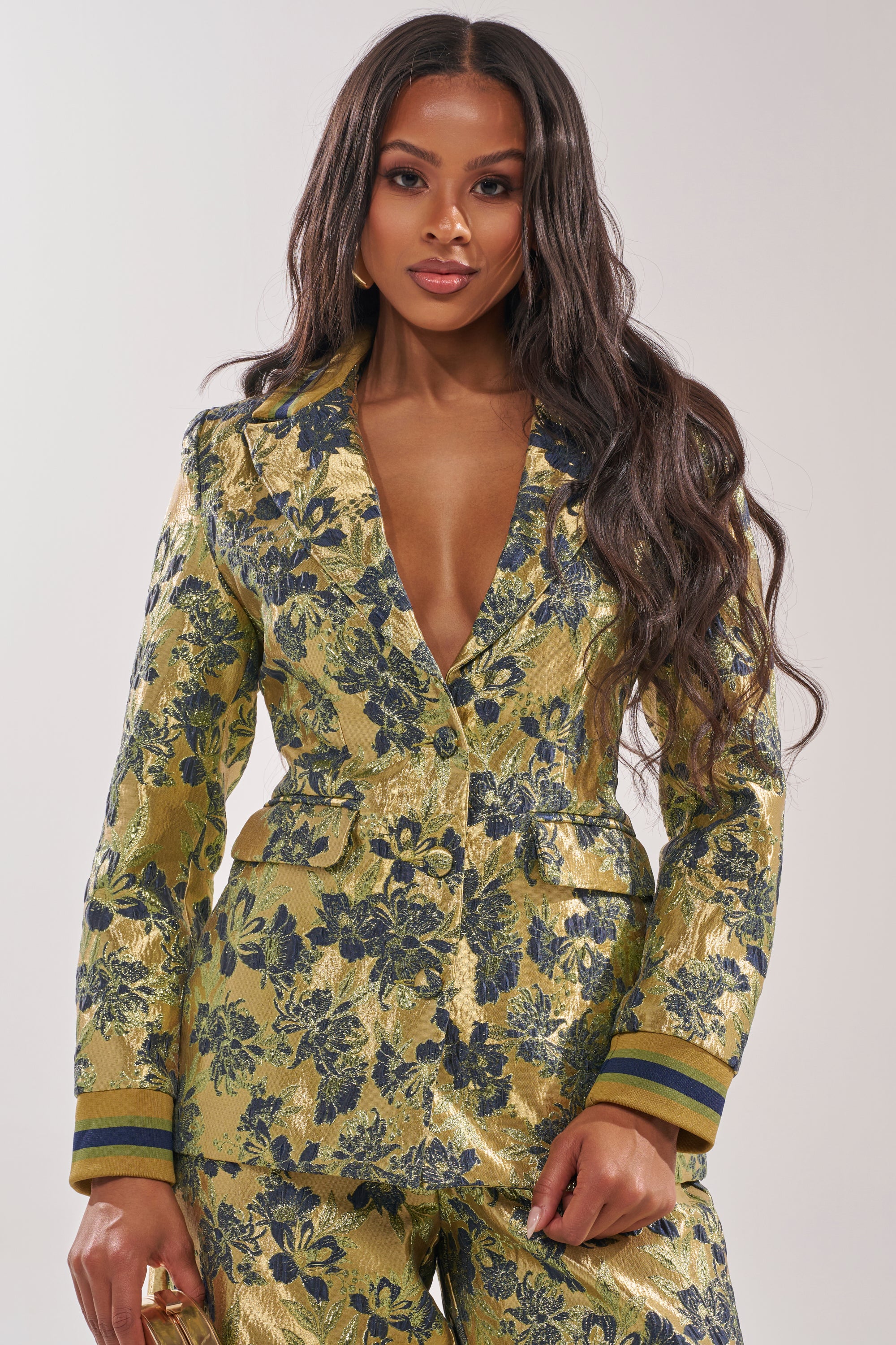 A woman with long, wavy hair confidently poses in the HARLOW BROCADE BLAZER, featuring metallic brocade fabric, blue embroidered floral details, and striped cuffs against a plain background. 0