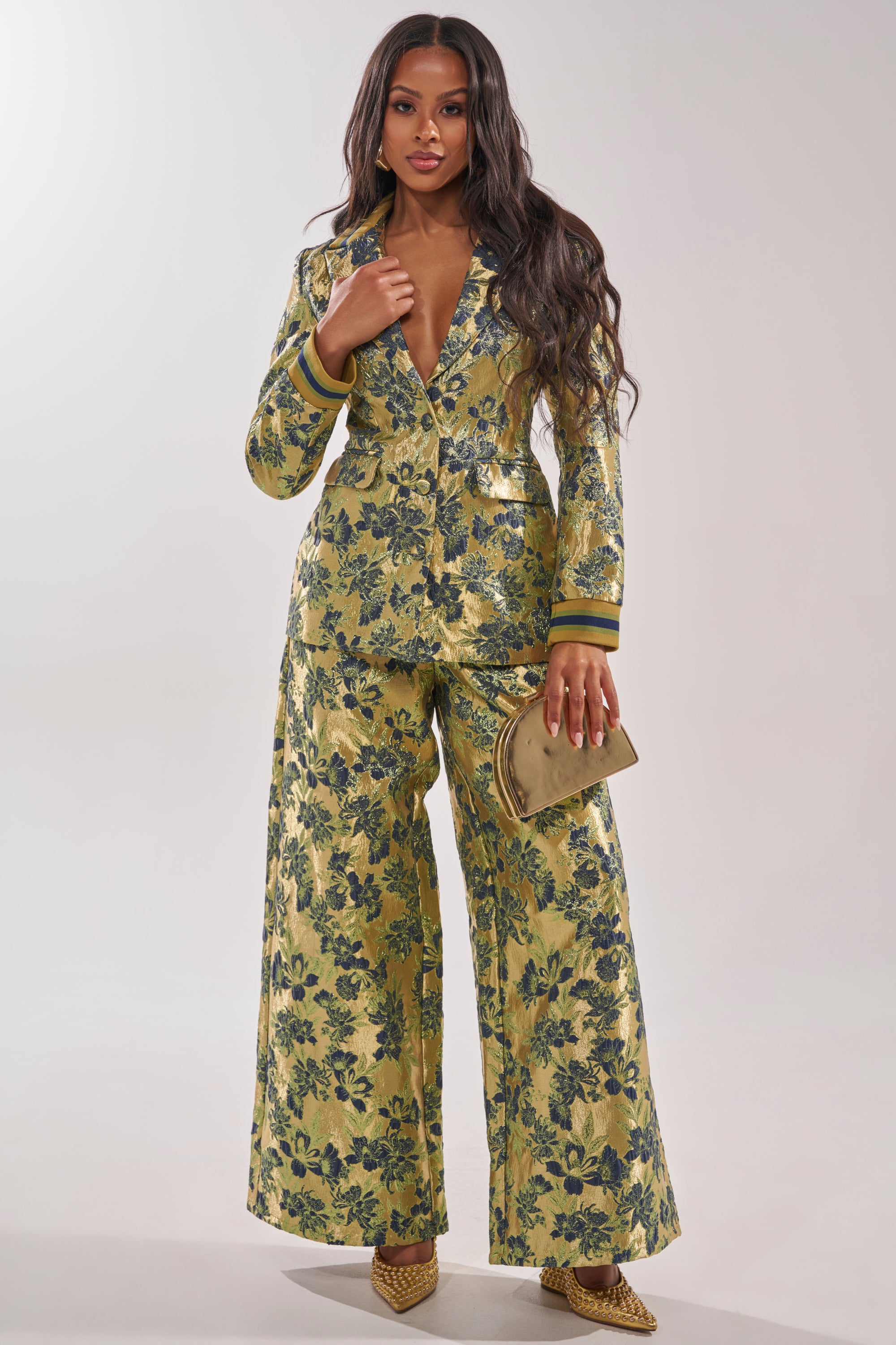 A woman wears the HARLOW BROCADE BLAZER, a shiny gold, deep V-neck metallic blazer with embroidered floral details, styled with wide-leg trousers, a metallic gold clutch, and pointed gold heels. 8