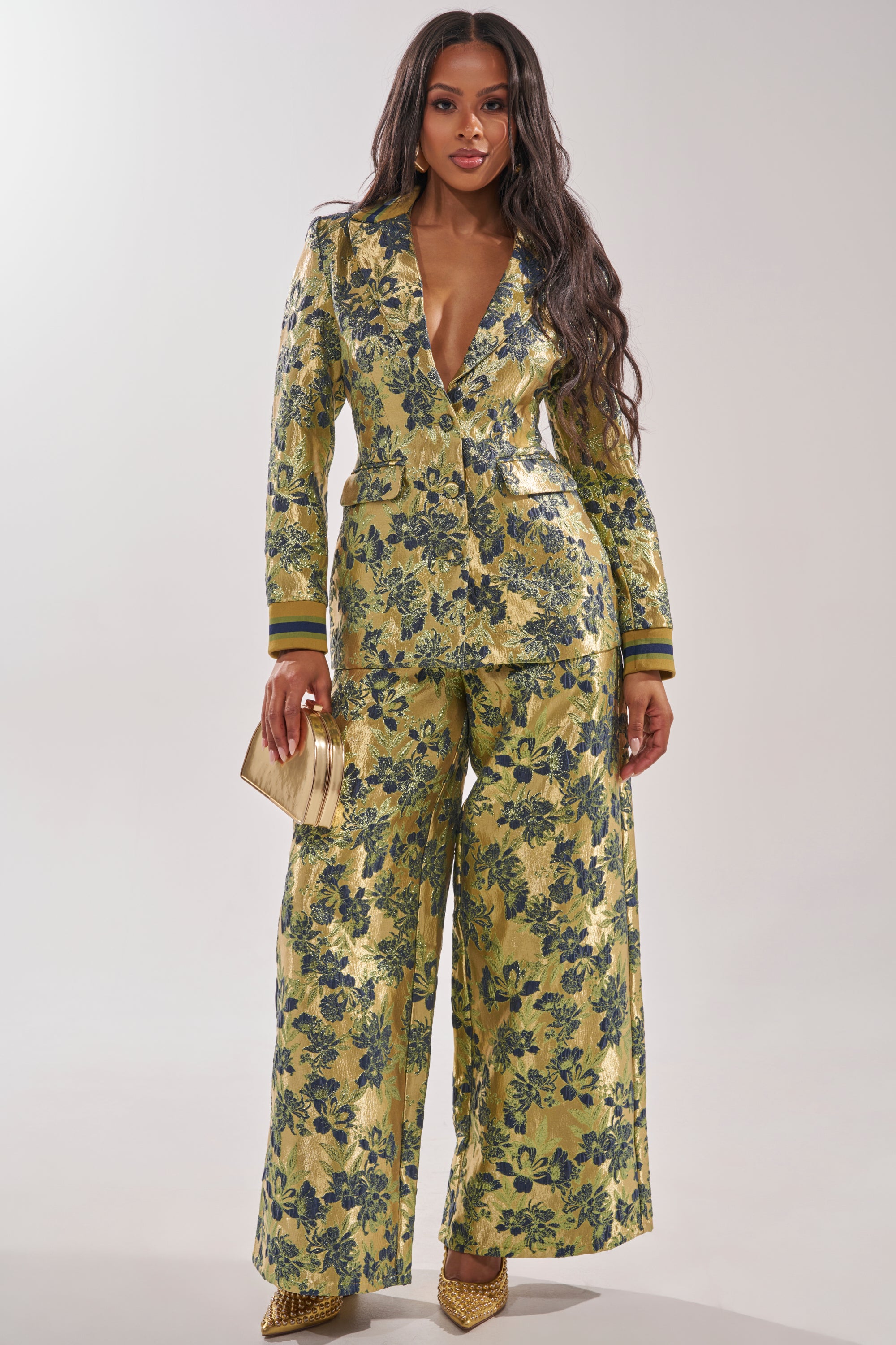 Wearing the HARLOW BROCADE BLAZER with blue floral embroidery, a woman stands confidently in matching gold suit and wide-leg pants, holding a gold clutch and wearing gold heels, her long wavy hair cascading over her shoulders. 5