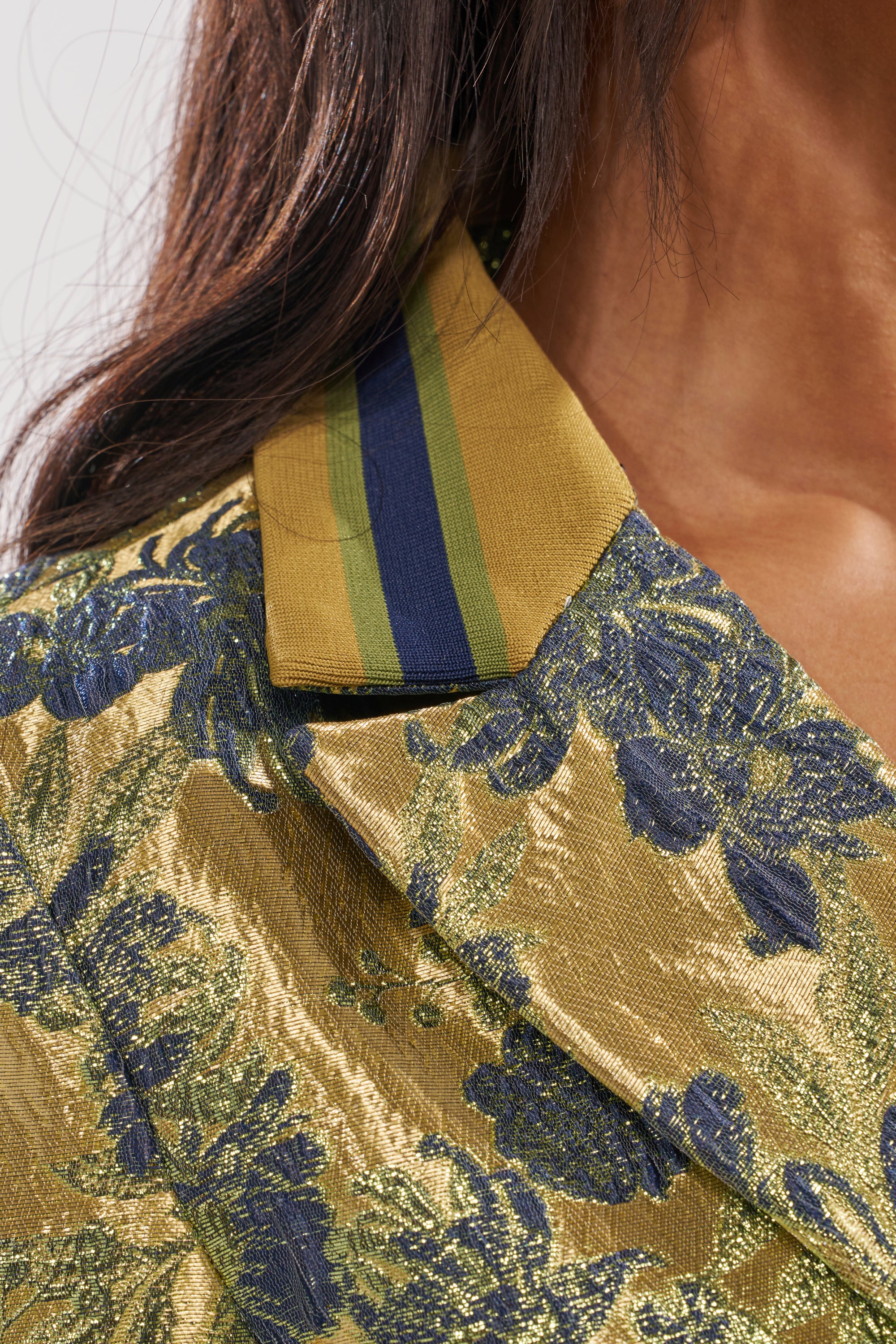Close-up of a person wearing the HARLOW BROCADE BLAZER in metallic gold and blue with embroidered floral details. The wide collar features olive green, navy blue, and gold stripes; only part of the face and shoulder are visible. 7