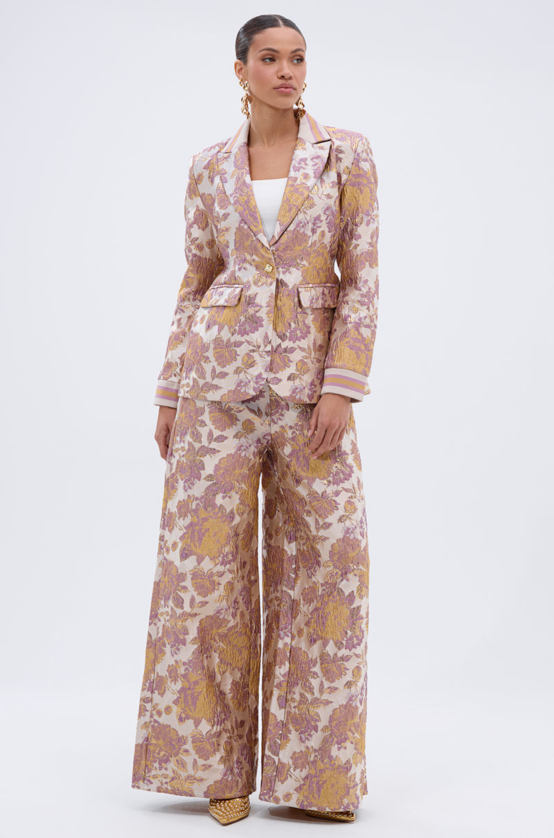 A woman wears the SLOW MOTION BROCADE BLAZER, featuring a pink and gold floral pattern, paired with wide-leg trousers and a white top. She styles her hair back and accessorizes with gold earrings and heels.