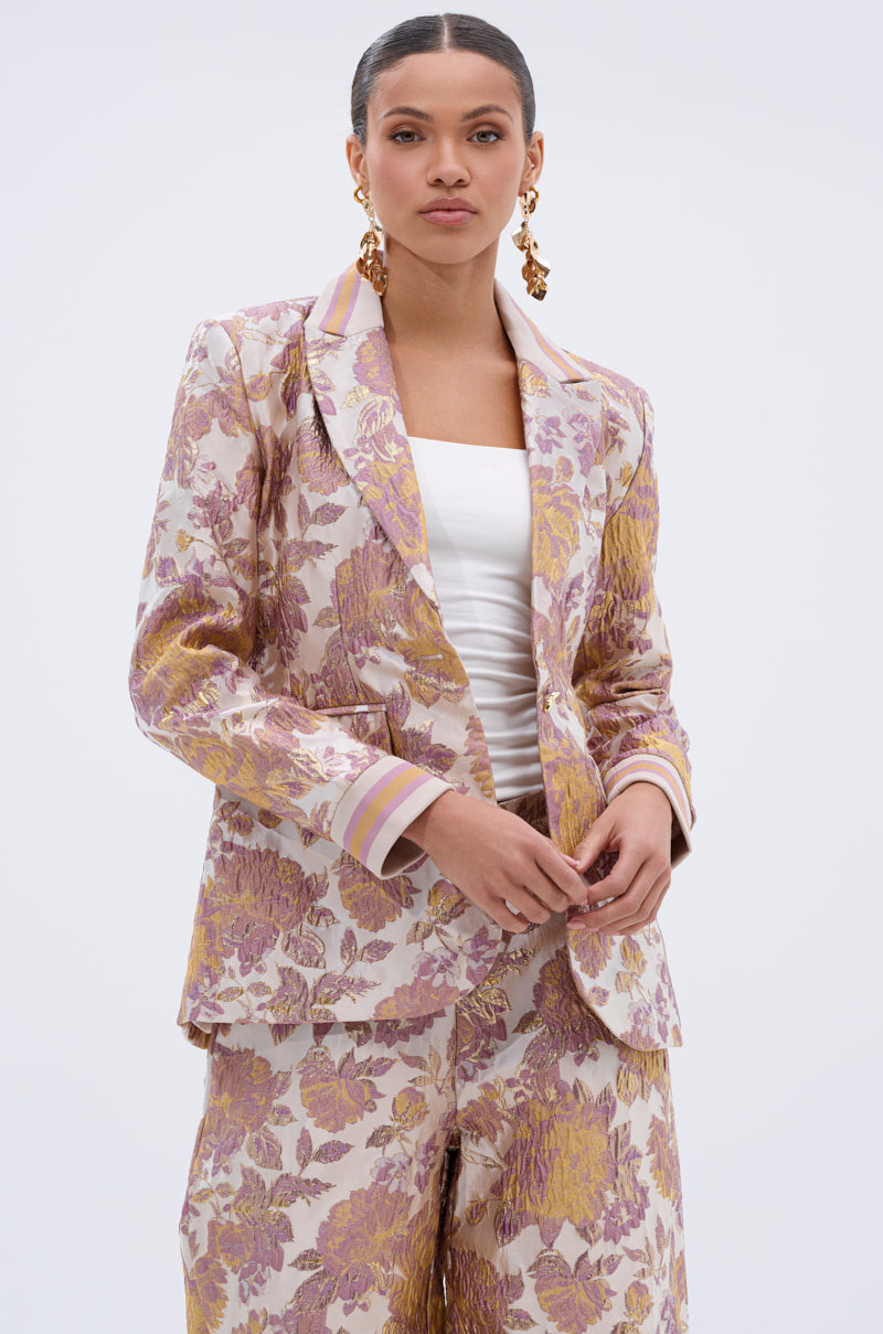 A woman stands against a plain light background wearing the SLOW MOTION BROCADE BLAZER in pastel floral patterns of purple and gold over a white top, paired with gold statement earrings and her hair pulled back.