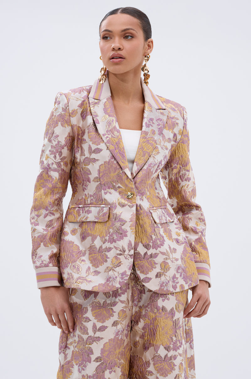 A woman poses in the SLOW MOTION BROCADE BLAZER—a pastel floral pattern blazer with gold and lavender metallic details, tailored fit, and wide-leg trousers. She accessorizes with gold earrings and has her hair pulled back against a plain background.