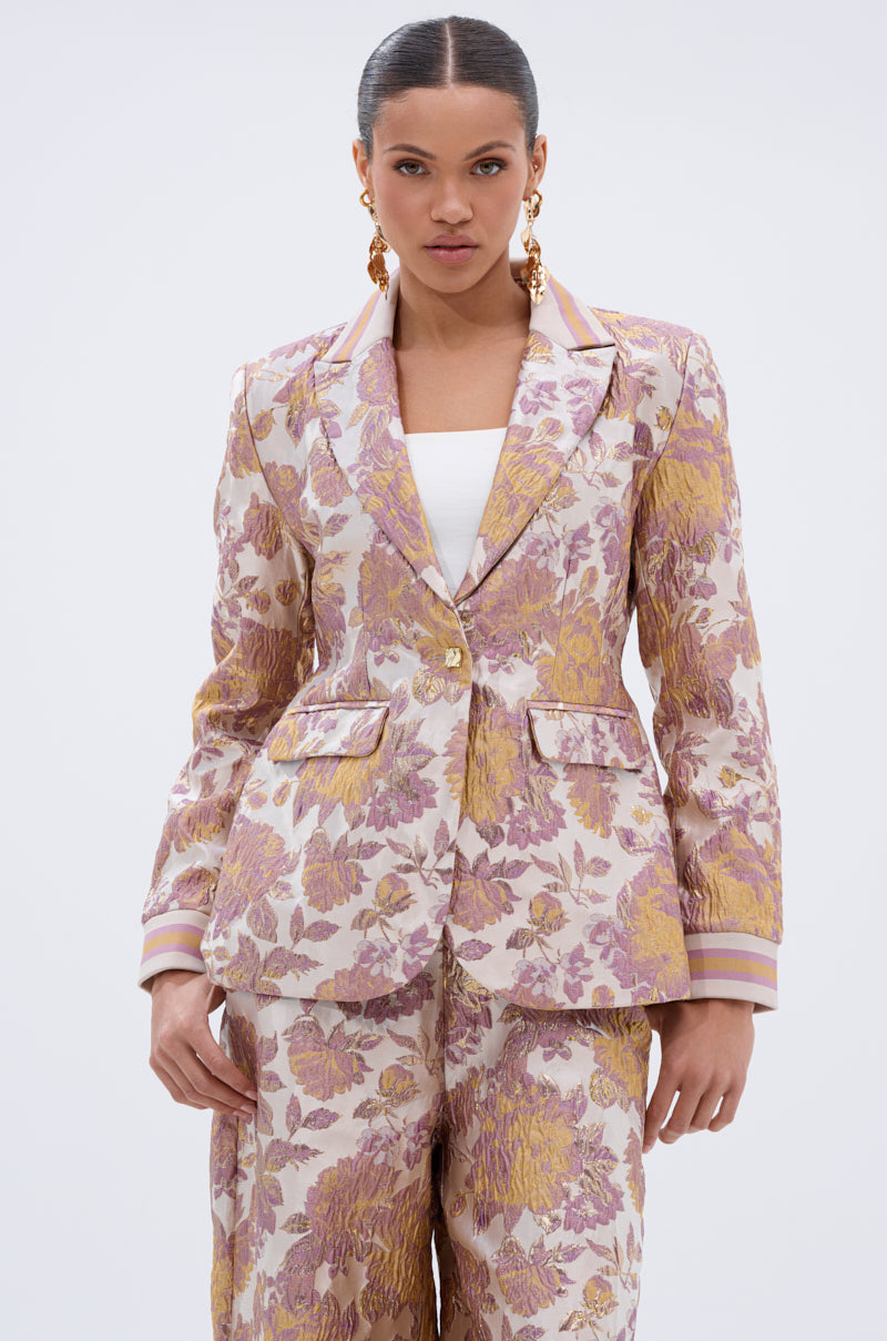 A woman stands against a plain background wearing the SLOW MOTION BROCADE BLAZER with gold, pink, and purple floral patterns. Her hair is pulled back, and she accessorizes with large gold statement earrings.