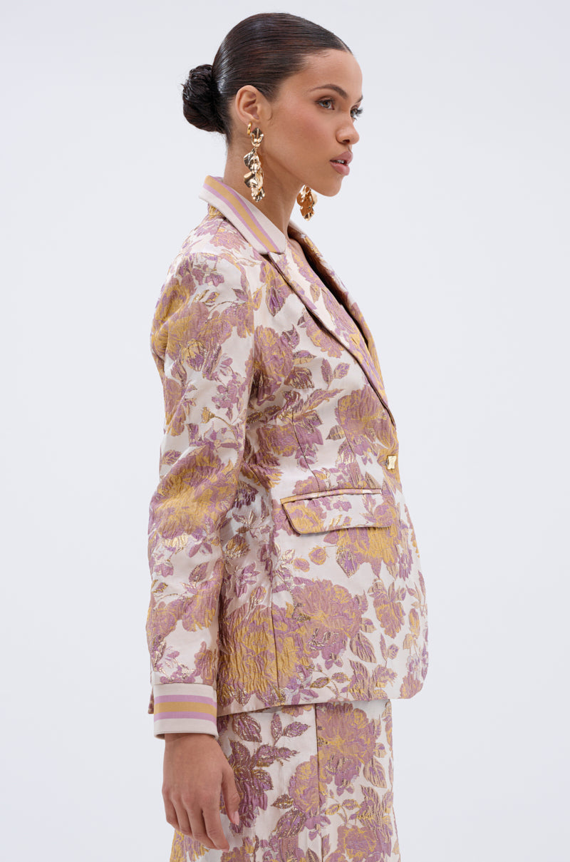 A woman with slicked-back hair models the SLOW MOTION BROCADE BLAZER in pink, gold, and cream with large gold earrings, posing in profile against a plain white background.