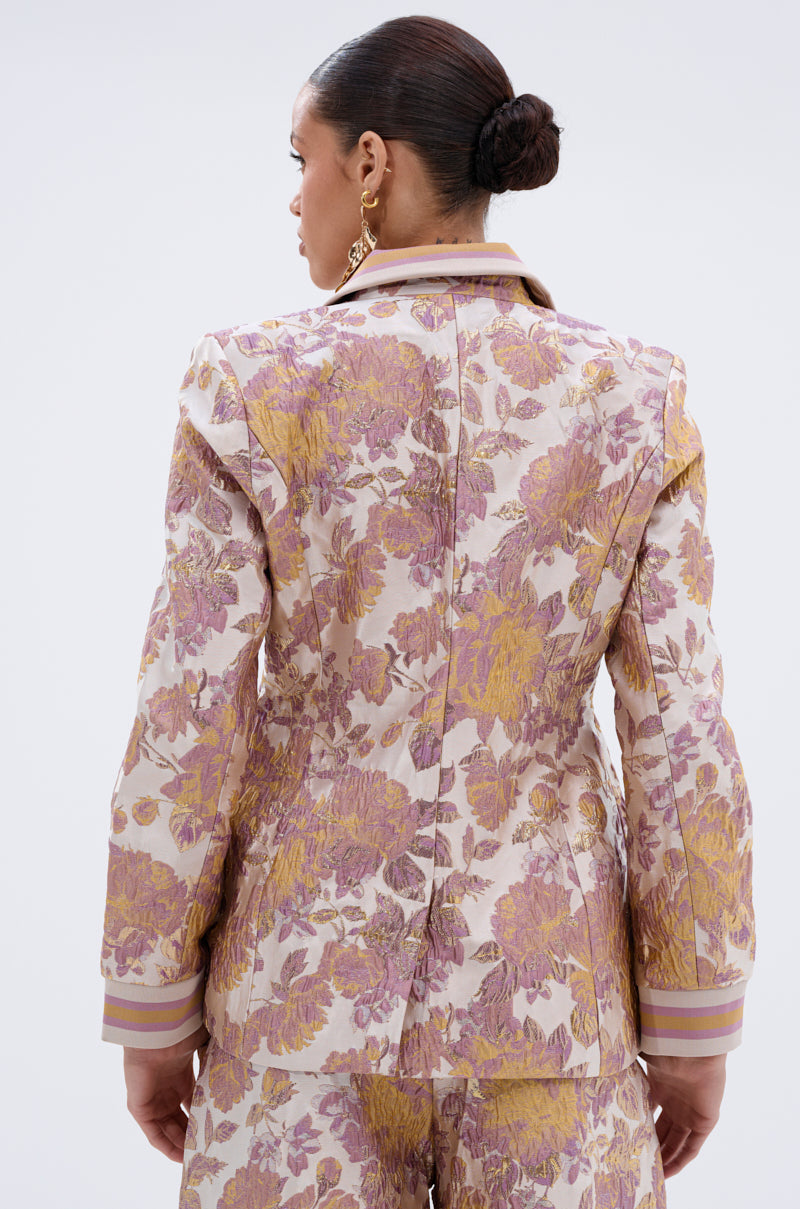 A woman with her hair in a low bun is seen from the back wearing the SLOW MOTION BROCADE BLAZER, featuring a fitted silhouette with a floral brocade pattern in purple, gold, and cream. She accessorizes with dangling earrings.