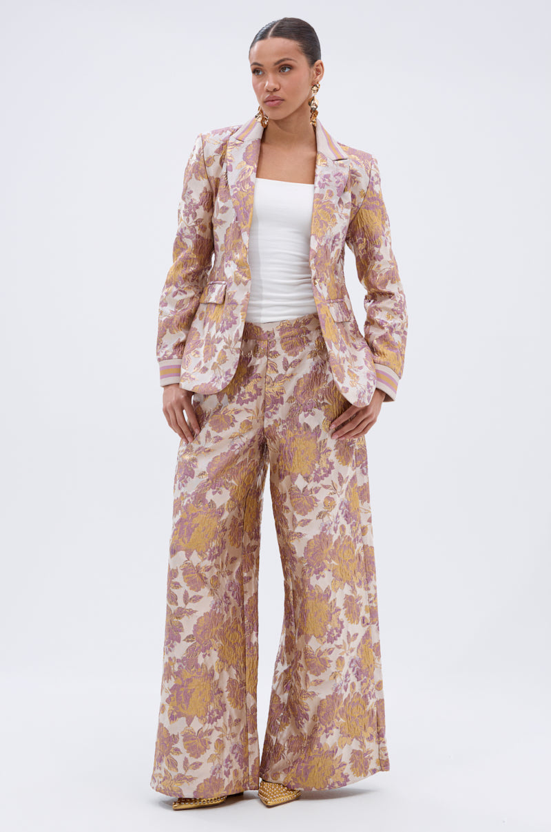 A woman stands facing forward in the SLOW MOTION BROCADE BLAZER—a pink, gold, and cream blazer—paired with wide-leg trousers, a white tube top, statement earrings, and pointed yellow shoes against a plain background.