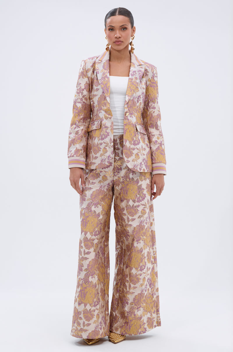 A woman poses against a plain background wearing the SLOW MOTION BROCADE BLAZER in pink and gold, paired with flared pants, a white top, statement earrings, and gold pointed shoes.