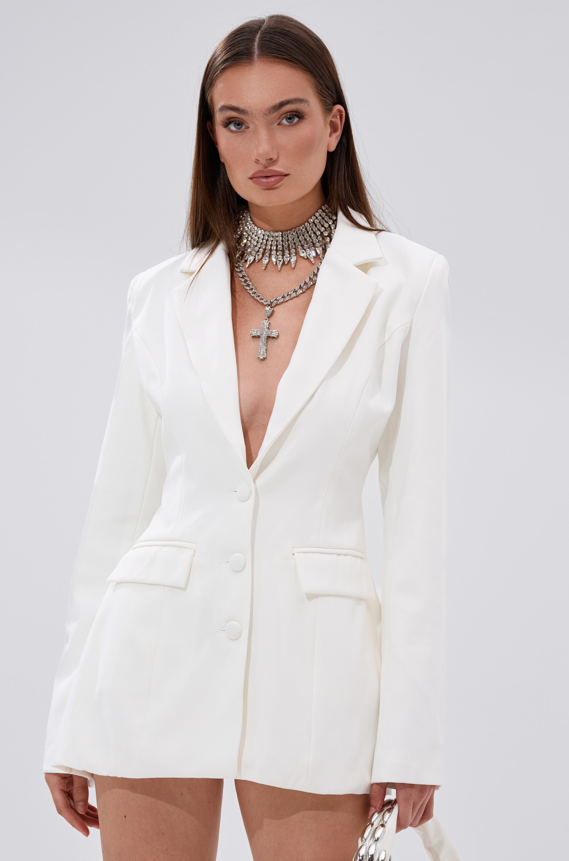 A woman with straight brown hair and a neutral expression models the EASY BREEZY BACKLESS BLAZER with no shirt underneath, styled with layered silver necklaces and a bold cross pendant, against a plain light background. 3