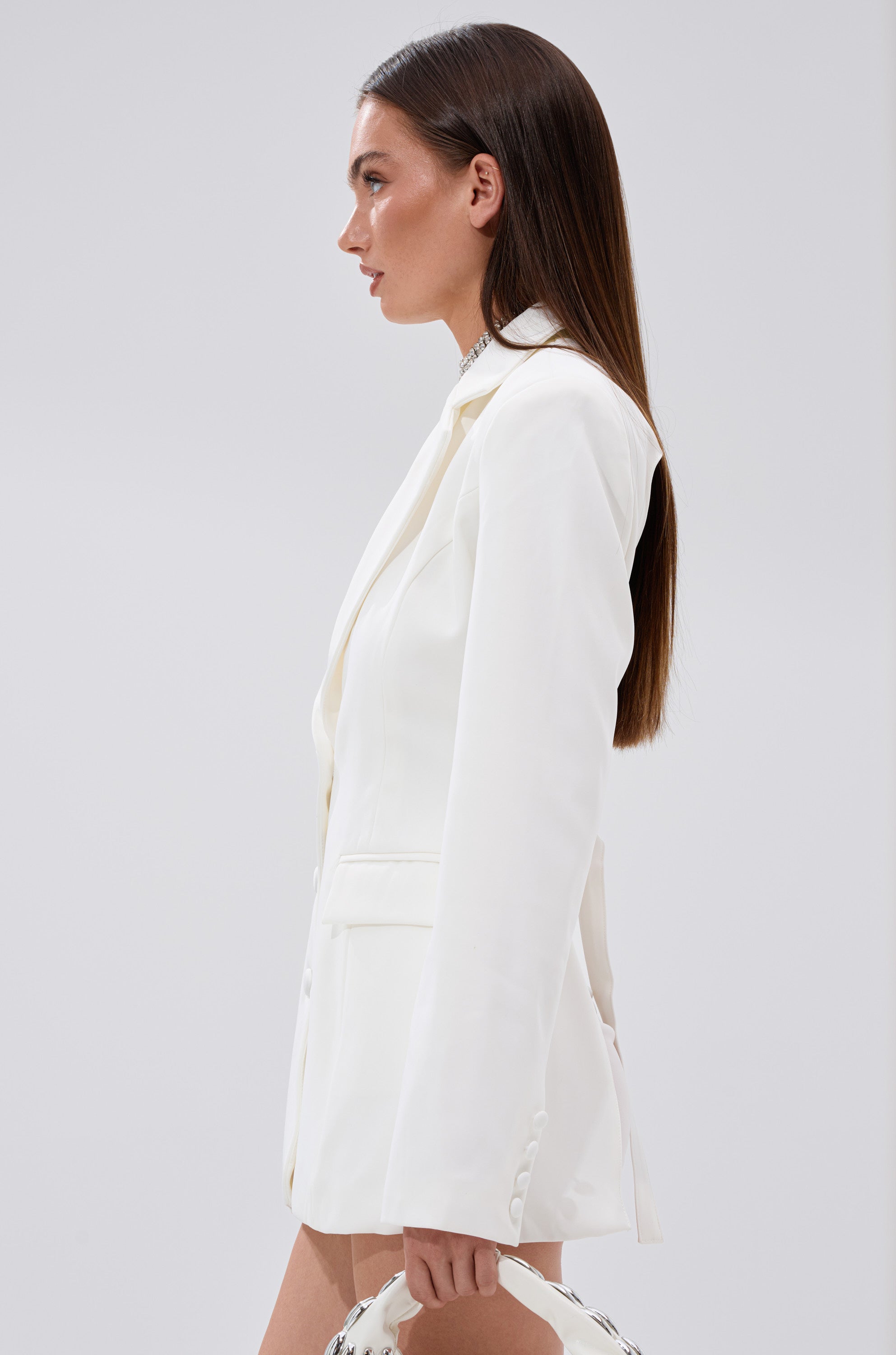 A woman with long straight brown hair is seen in profile wearing the EASY BREEZY BACKLESS BLAZER and holding a white handbag against a plain light background. 4