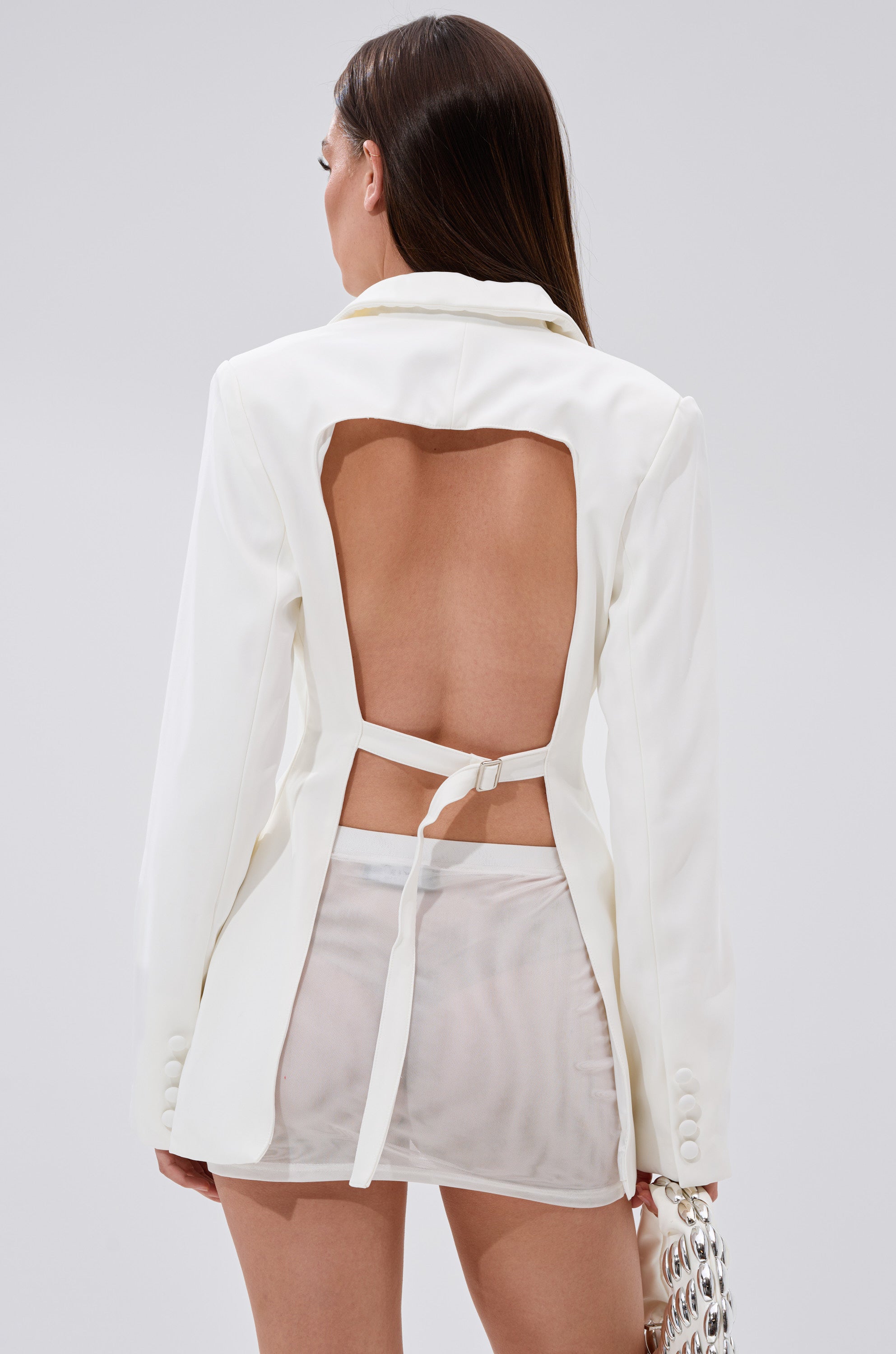 A woman with long brown hair models the EASY BREEZY BACKLESS BLAZER, a white blazer with strap detail, paired with a sheer mini skirt. She holds a metallic handbag and stands against a simple, light background. 0