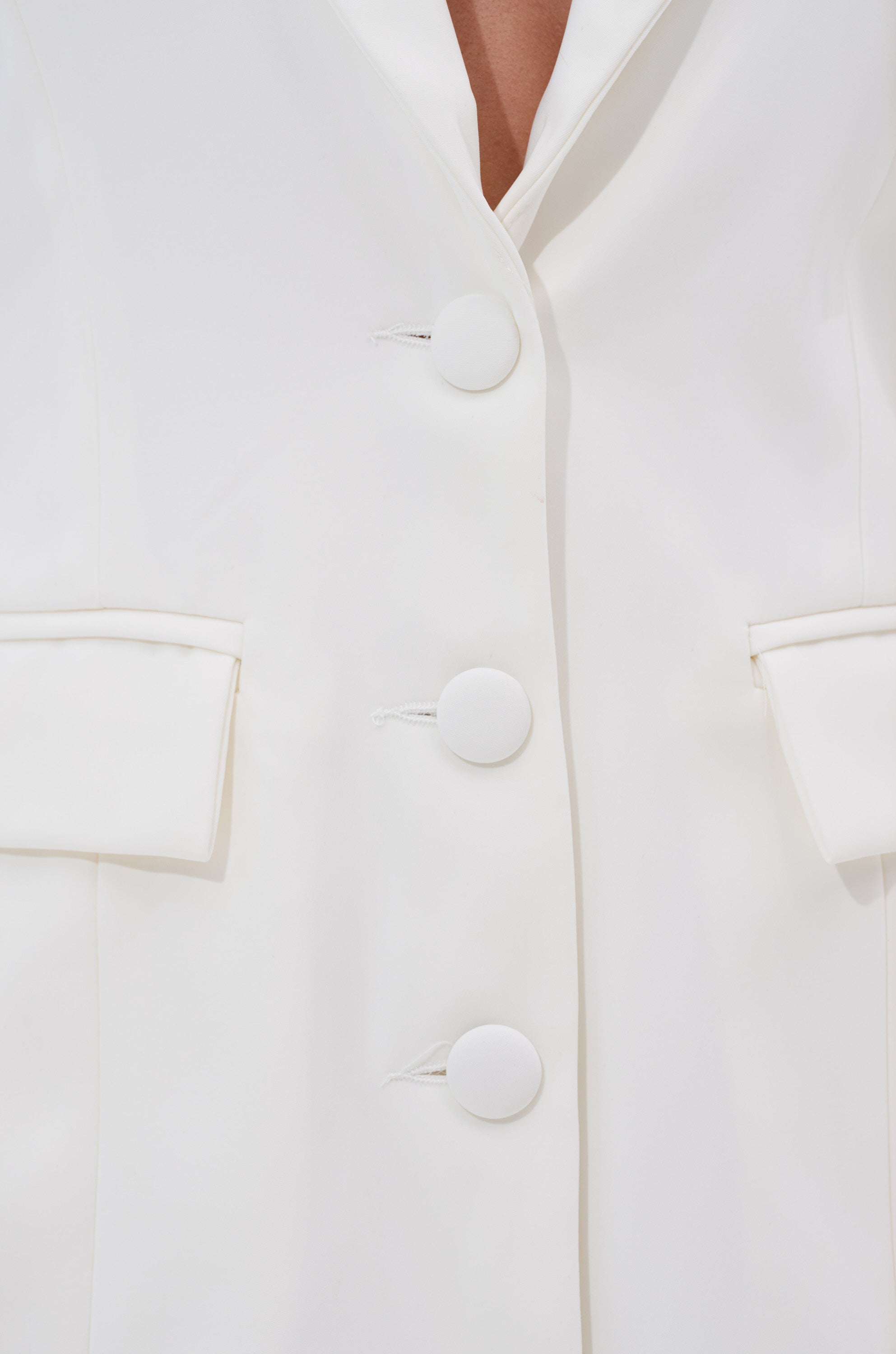 Close-up of the EASY BREEZY BACKLESS BLAZER—a tailored white blazer featuring large matching buttons, two flap pockets, and an open collar that reveals a glimpse of the wearer's chest. 6