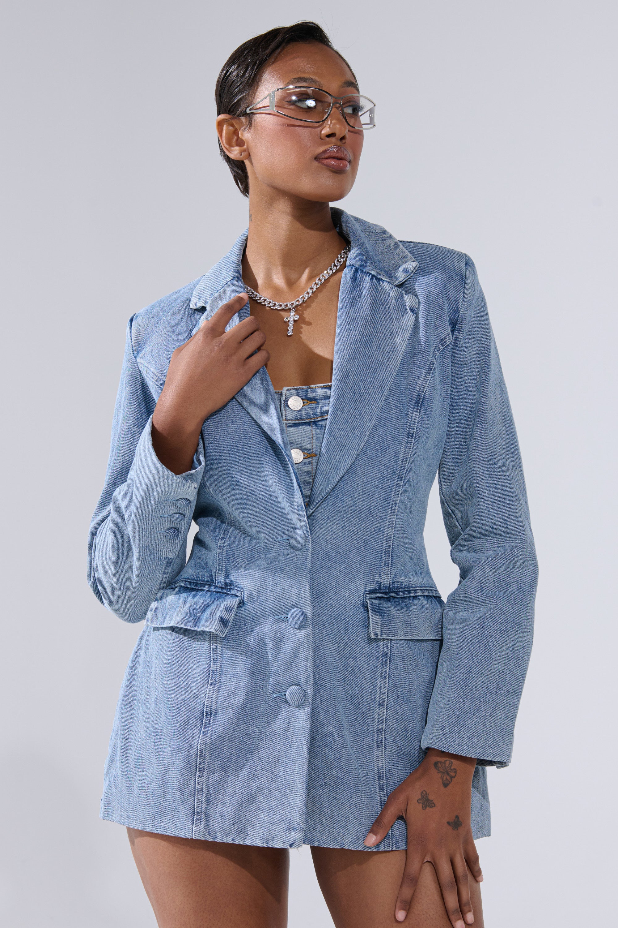 A person models the BACKLESS DENIM BLAZER—light-wash and matched with a similar top—styled with clear glasses and a silver chain necklace, posing confidently against a plain light background.
