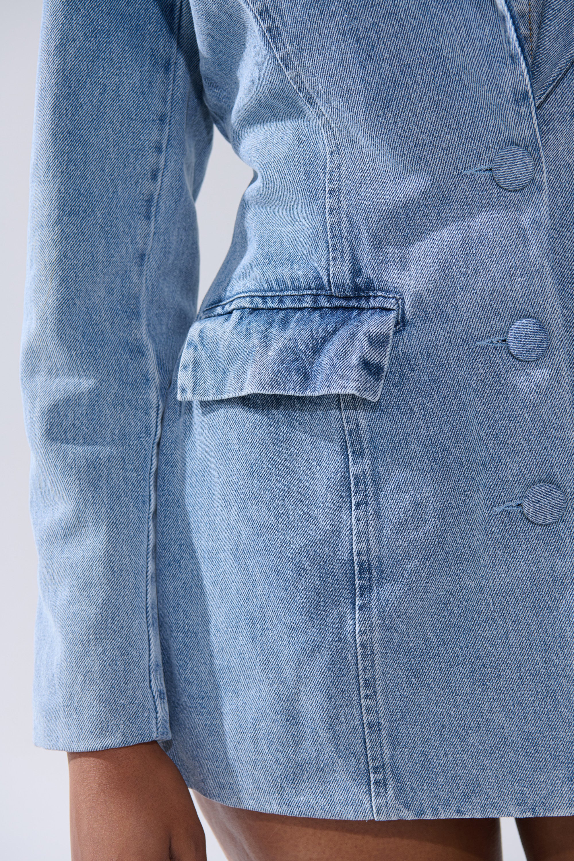 Close-up of a person wearing the BACKLESS DENIM BLAZER, highlighting the front buttons, flap pocket, and part of their hand against a neutral background.
