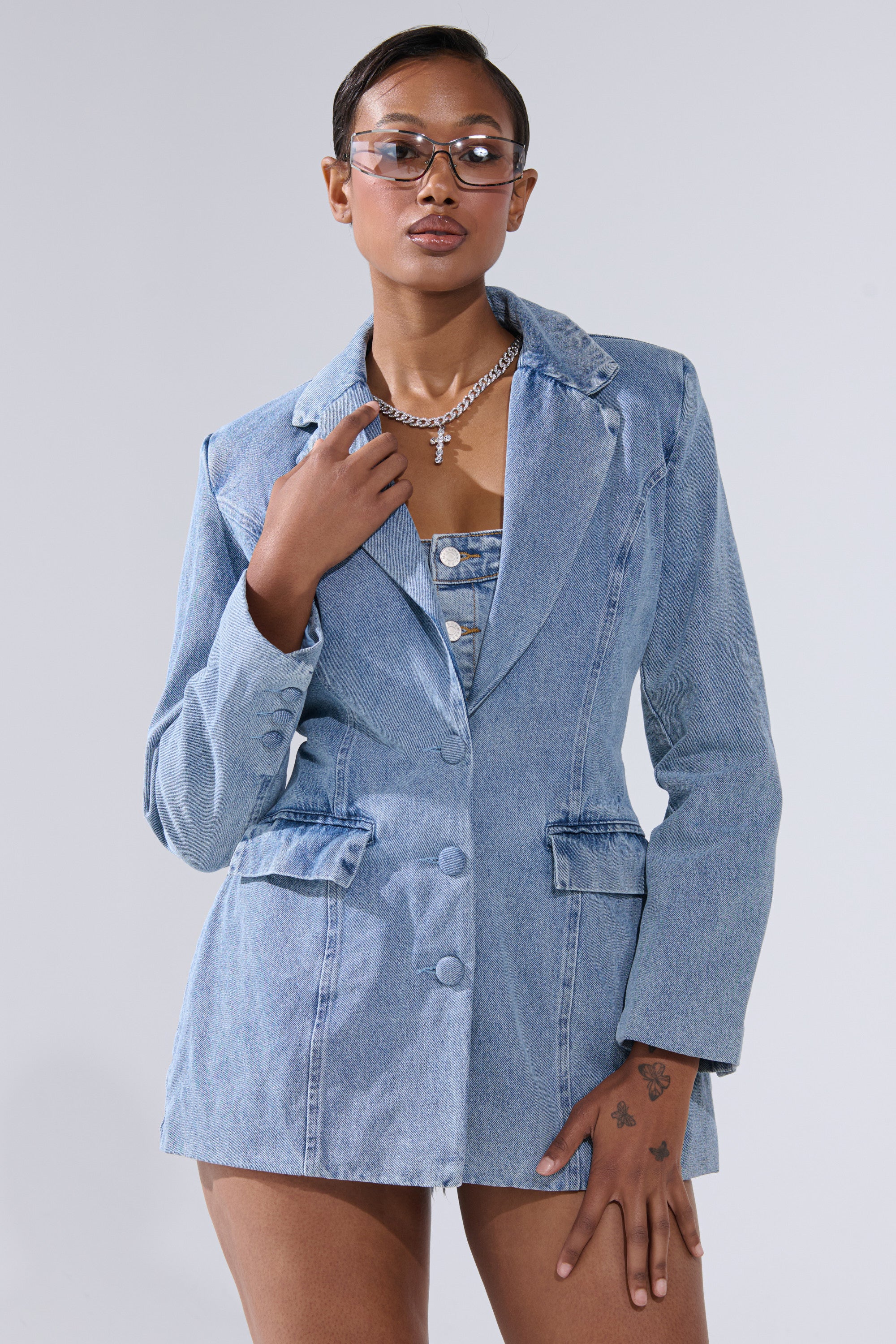 A woman poses confidently against a light gray background, wearing clear glasses, a silver necklace, and the BACKLESS DENIM BLAZER over a matching denim top, with one hand on her chest.