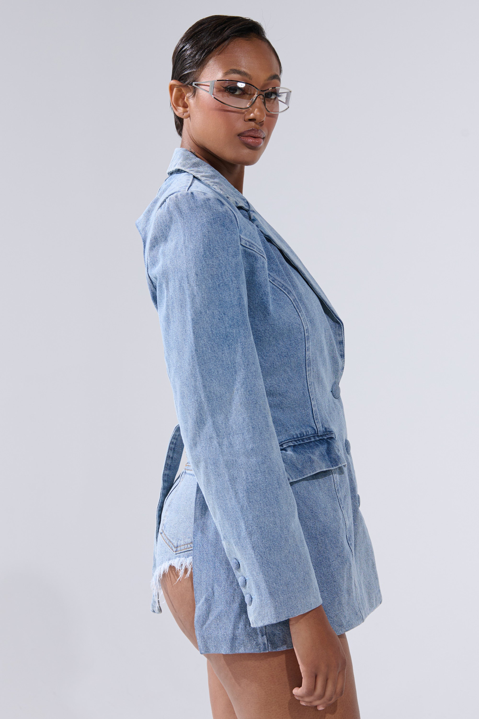 A woman in profile wears the BACKLESS DENIM BLAZER with matching high-waisted, frayed denim shorts. She has slicked-back hair, stylish narrow glasses, and stands against a plain, light background.