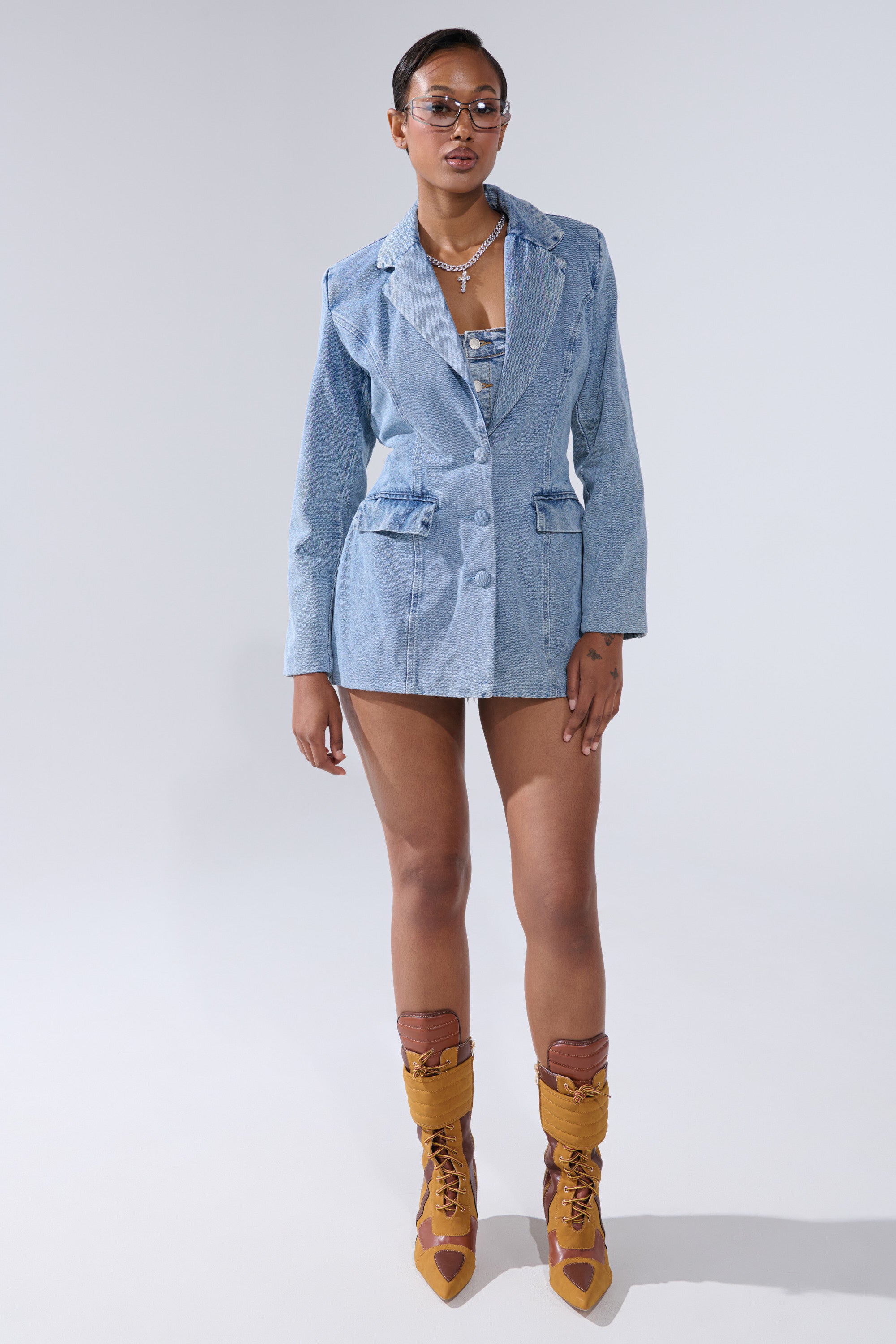 A woman poses against a plain backdrop in the BACKLESS DENIM BLAZER, styled with a matching top and tan lace-up heeled boots. She completes her look with layered necklaces, earrings, and glasses.