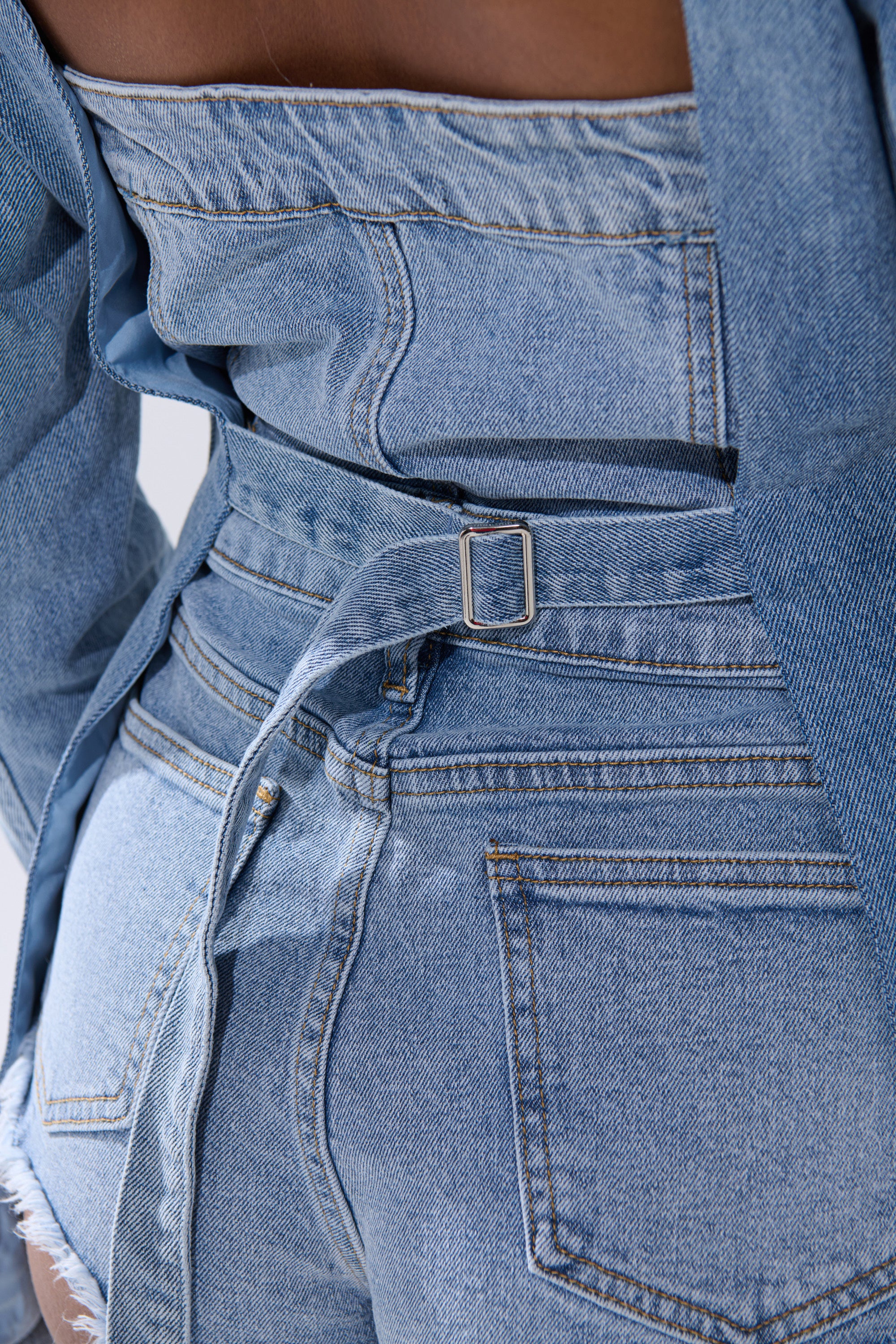 Close-up of a person wearing the BACKLESS DENIM BLAZER styled as a belted jacket with high-waisted denim shorts. Visible stitching, back pockets, and textured seams highlight this women’s denim look.