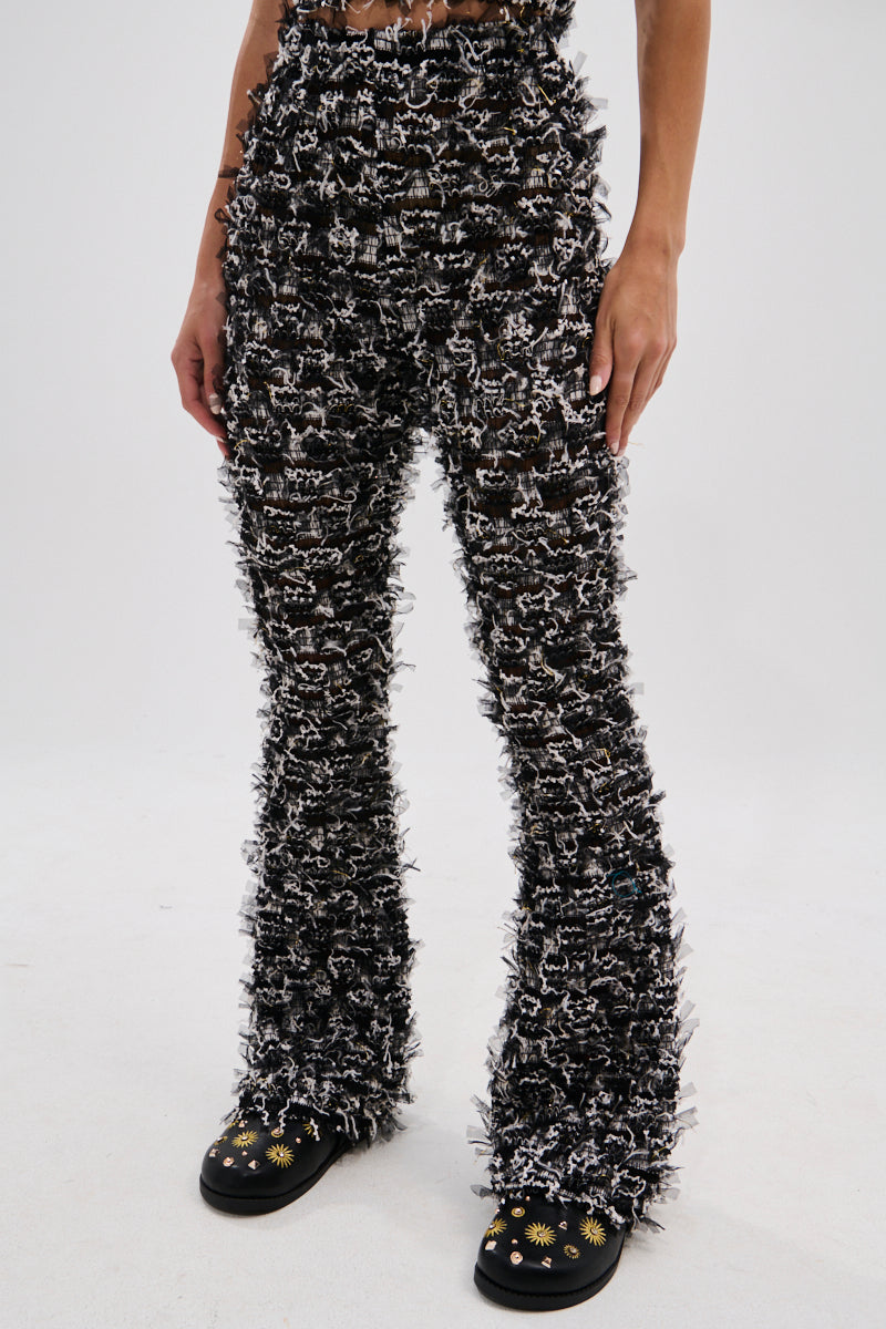 Wearing the CRAZY FOR YOU FRINGE FLARE LEGGING in textured black and white with a high-rise fit and fringe flare style, paired with black shoes adorned with yellow and white floral designs—perfect for music festivals against a plain background. 1