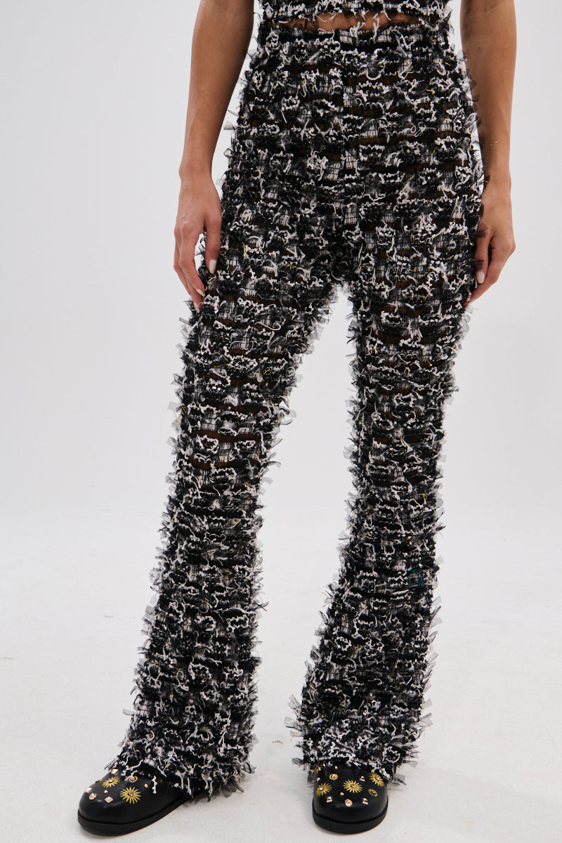 A person wears the CRAZY FOR YOU FRINGE FLARE LEGGING—high-rise, black and white textured fuzzy fringe flares—paired with black shoes featuring yellow and white flower designs, perfect for music festivals, on a plain white background. 2