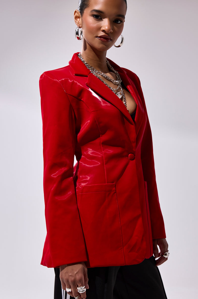 Wearing the TOO SLICK LIQUID VELVET BLAZER in shiny red along with layered silver chain necklaces, hoop earrings, and rings, a person confidently poses against a plain light background.