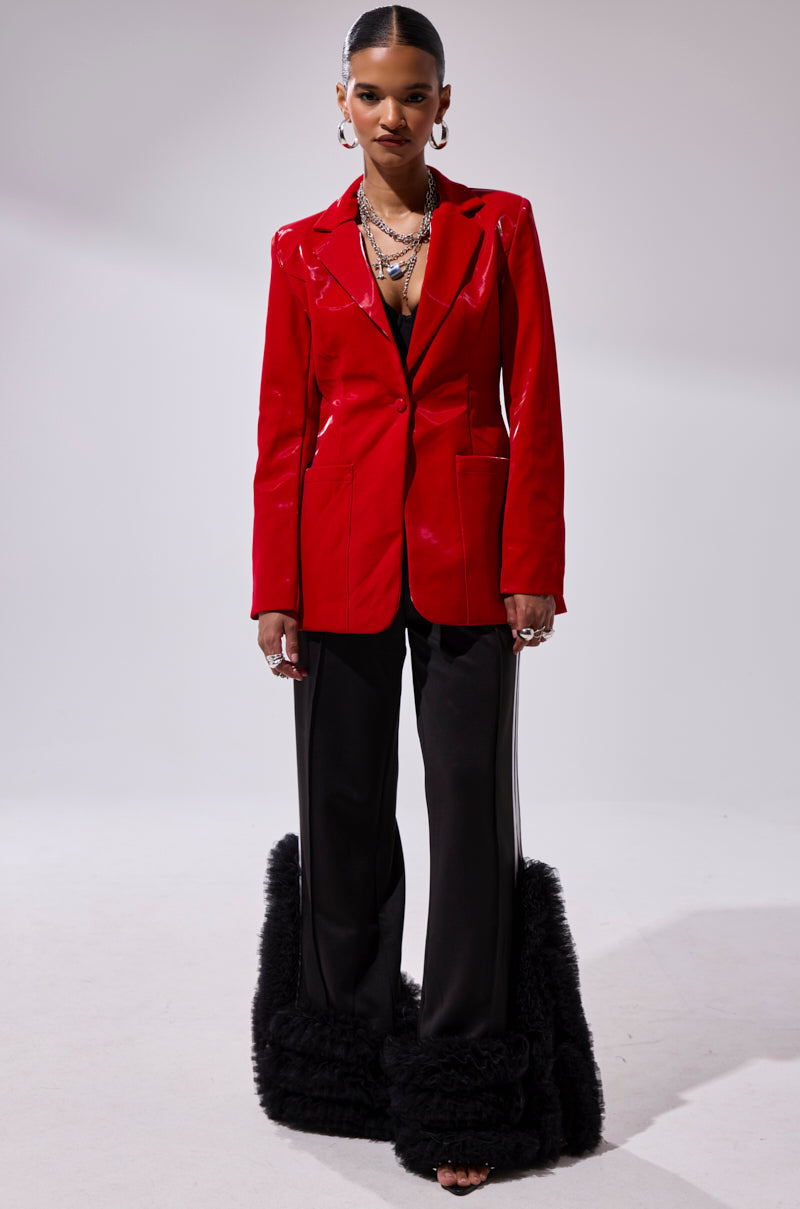 A woman poses against a plain light backdrop wearing the TOO SLICK LIQUID VELVET BLAZER in red faux patent leather over black wide-leg pants with large fluffy cuffs, accessorized with layered necklaces, hoop earrings, and slicked-back hair.