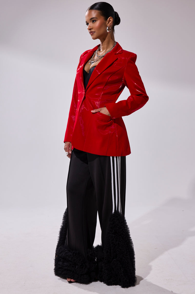 A woman in profile wears the TOO SLICK LIQUID VELVET BLAZER, paired with black wide-leg pants featuring white stripes and furry trim. She accessorizes with statement earrings, layered necklaces, and has slicked-back hair against a plain background.