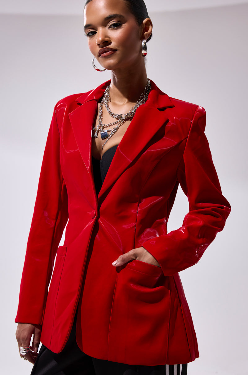A confident woman wears the TOO SLICK LIQUID VELVET BLAZER—shiny faux patent leather—with one hand in her pocket, layered necklaces, hoop earrings, and black clothing underneath, standing against a plain white background.