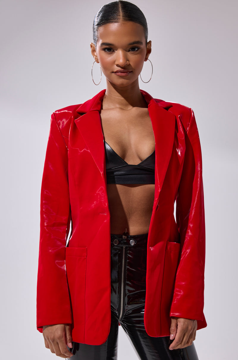 A woman with hoop earrings wears the TOO SLICK LIQUID VELVET BLAZER over a black bralette and glossy black pants, standing confidently against a plain background.