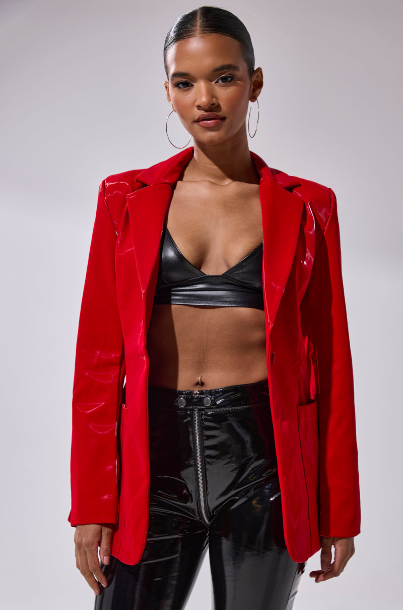 A woman stands confidently against a plain background, wearing the TOO SLICK LIQUID VELVET BLAZER in shiny red over a black bralette and black vinyl pants. She accessorizes with hoop earrings and has her hair pulled back.