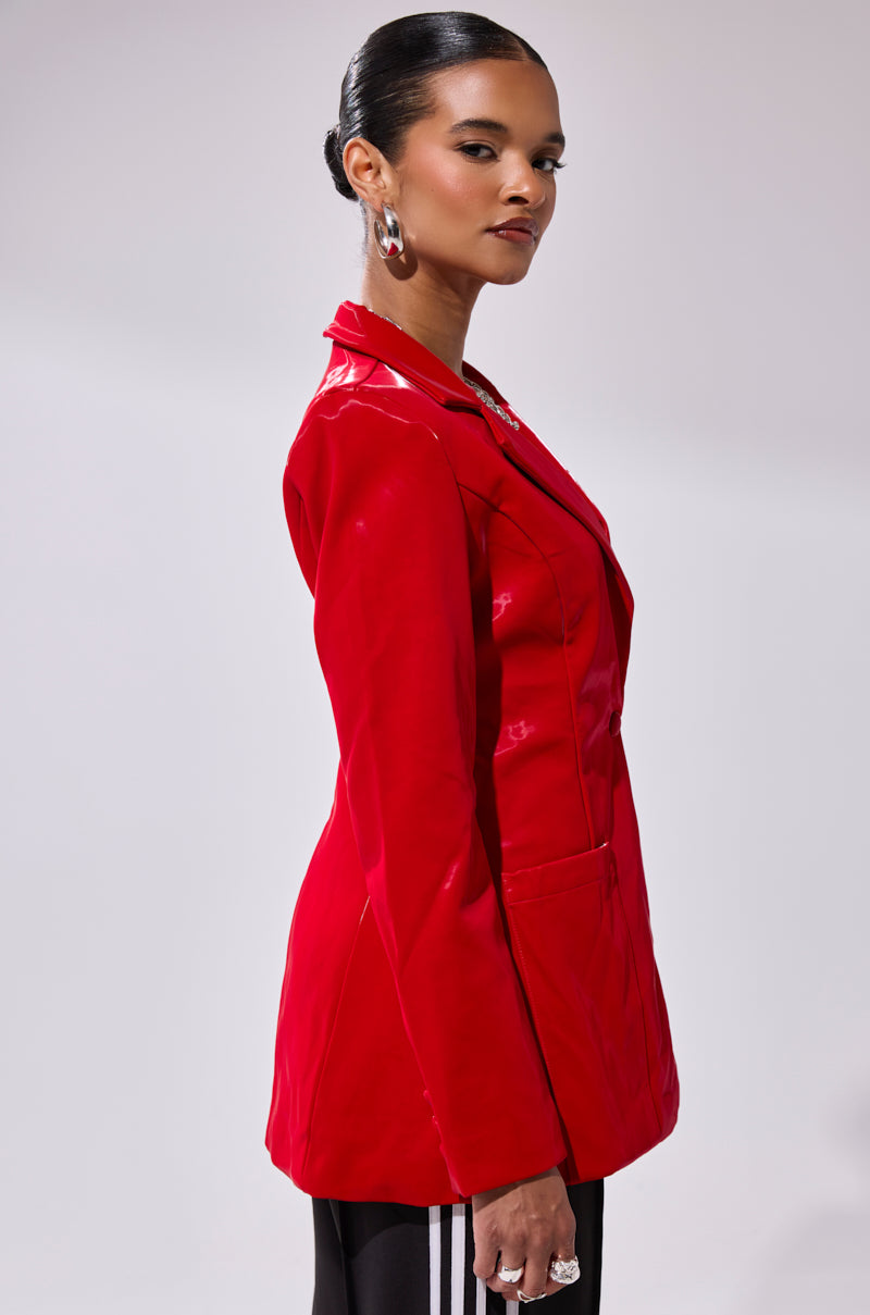 A woman with slicked-back hair wears the TOO SLICK LIQUID VELVET BLAZER, black striped pants, large hoop earrings, and rings, posing confidently in profile against a light background.