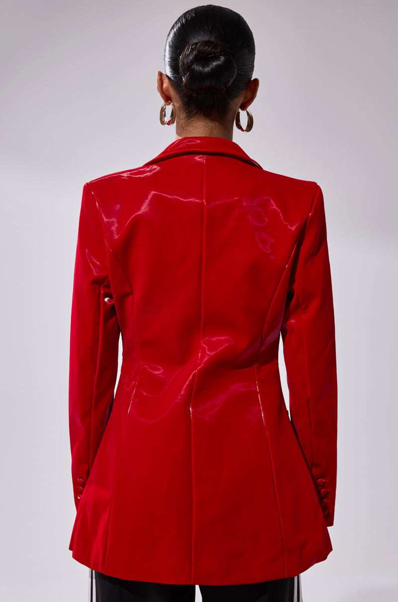 A person with their hair in a sleek bun, wearing large hoop earrings and the TOO SLICK LIQUID VELVET BLAZER in shiny red faux patent leather, is shown from the back against a plain light background.
