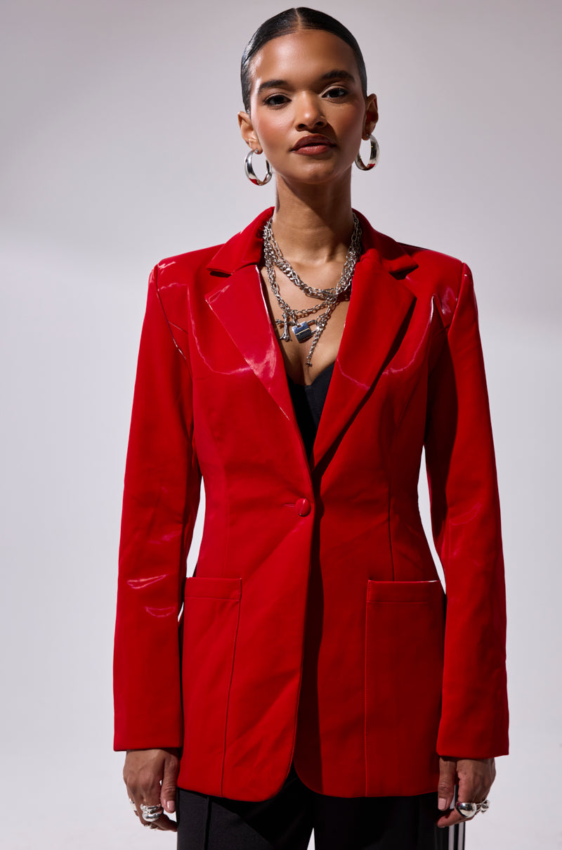 Someone models the TOO SLICK LIQUID VELVET BLAZER in bold red with shiny lapels, paired with layered silver necklaces, hoop earrings, and rings, posing against a simple light background.